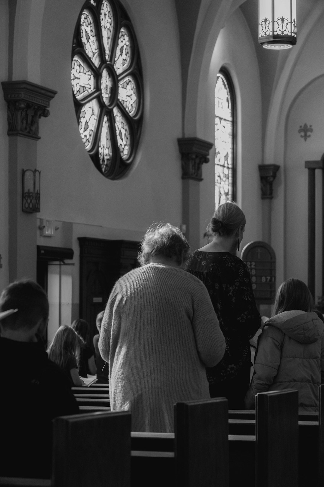 People praying inside a church with stained glass windows and wooden pews, captured in black and white.