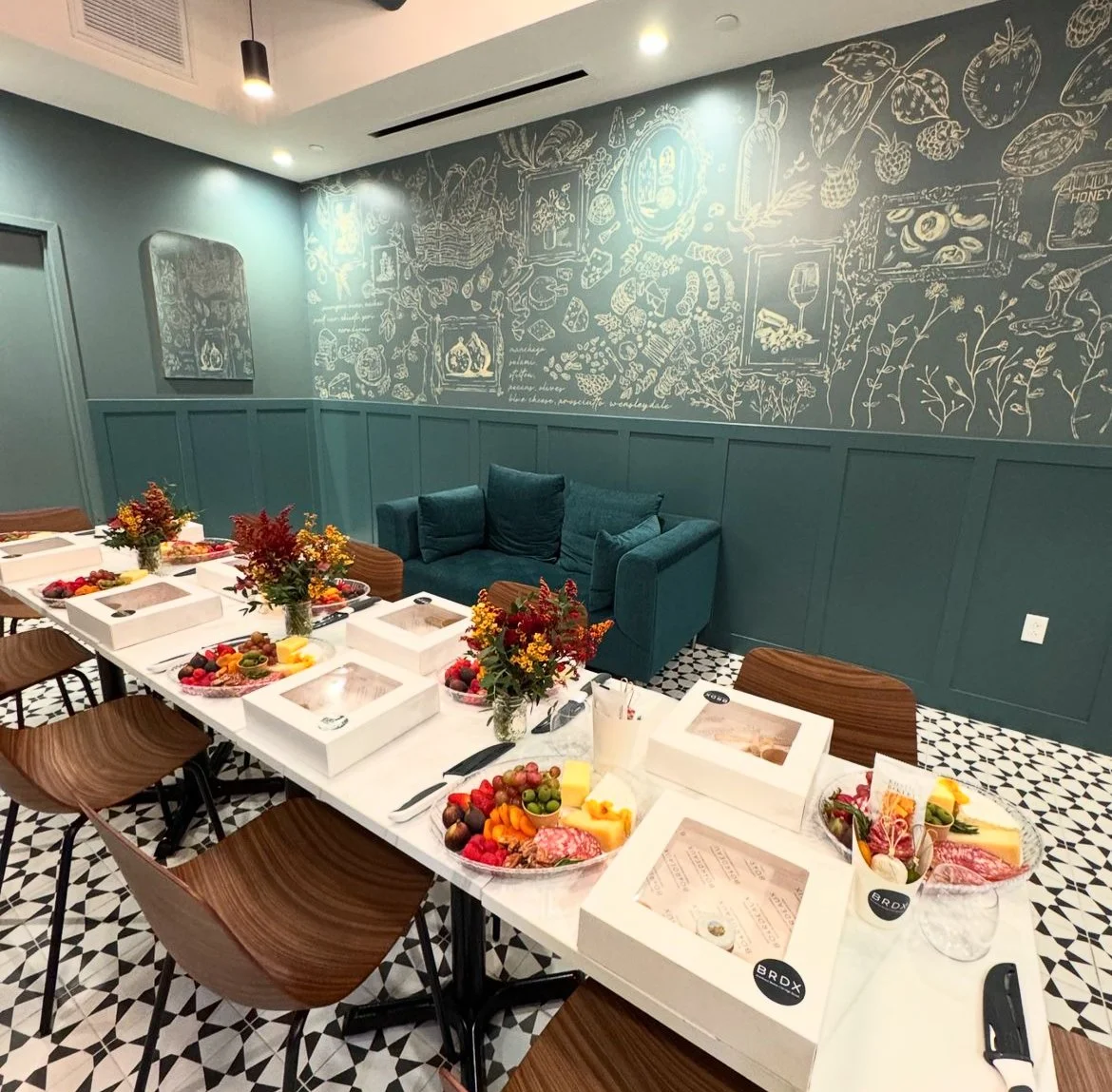 A dining room with a long table set for a meal, featuring cheese, fruit, and other food in white takeout boxes, surrounded by wooden chairs. The wall behind has teal panels and a chalkboard-style mural of various food and drink illustrations. There a