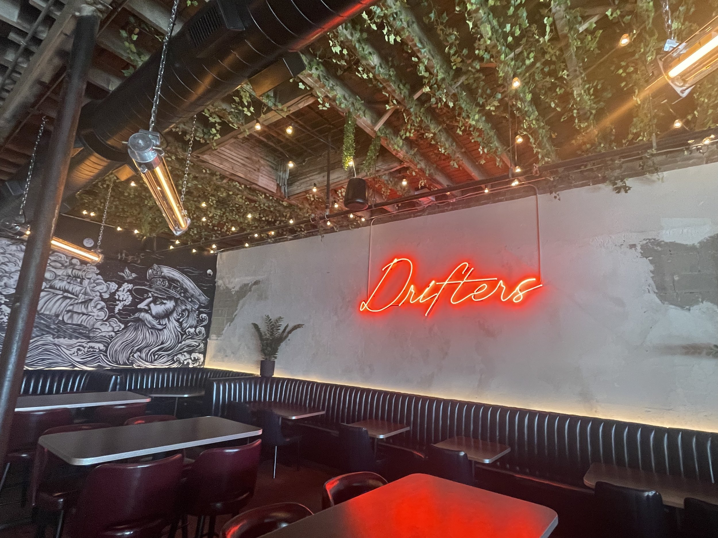 Restaurant with a black and white mural of a bearded sailor on the wall, a neon red sign that says 'Drifters,' booths, tables, and ceiling decorated with hanging greenery and string lights. Brand design by J Lenore Designs for Drifters, Quincy MA.