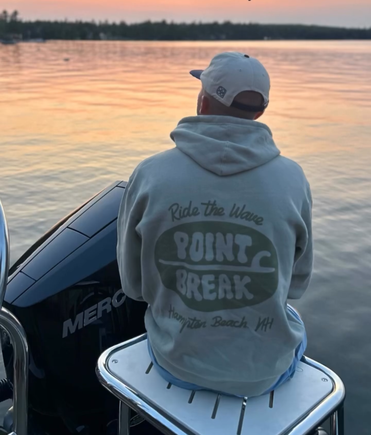 Person wearing a hoodie and cap sitting on a boat, looking at a calm body of water during sunset.