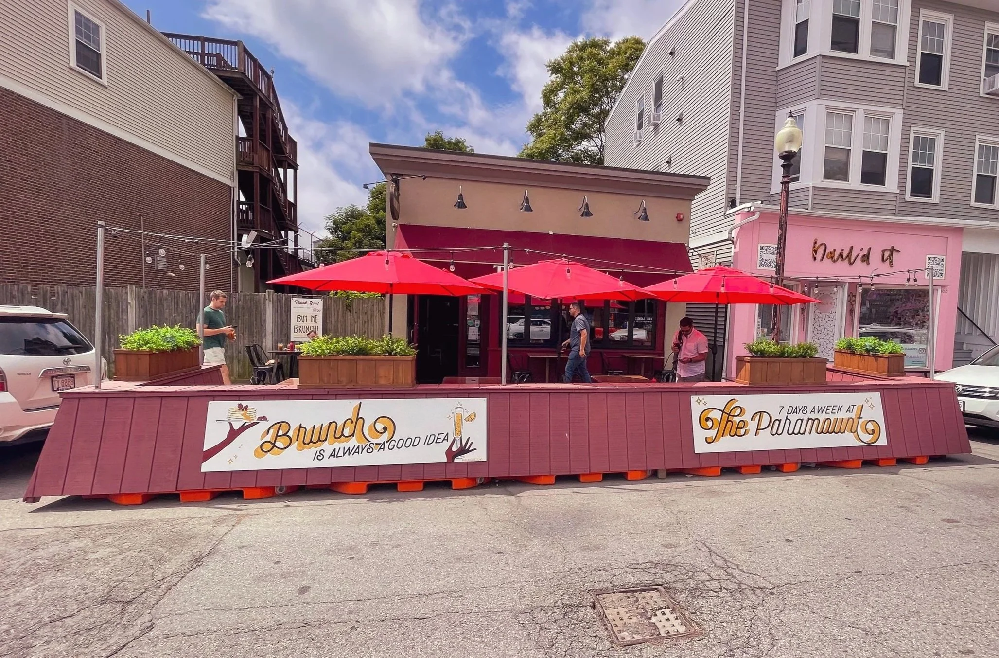 A pink building with a patio featuring red umbrellas and potted plants, with a sign that reads "Brunch is always a good idea" and "7 days a week at The Paramount." Custom signs for The Paramount, South Boston.