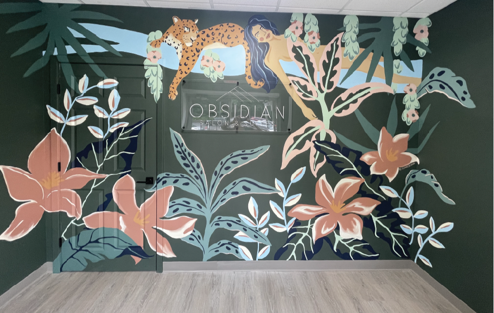 Colorful mural of a leopard lounging in a jungle scene with large pink flowers, leafy plants, and a woman with long dark hair laying among the foliage, with a sign that reads "Obsidian Salon & Beauty" in the background.