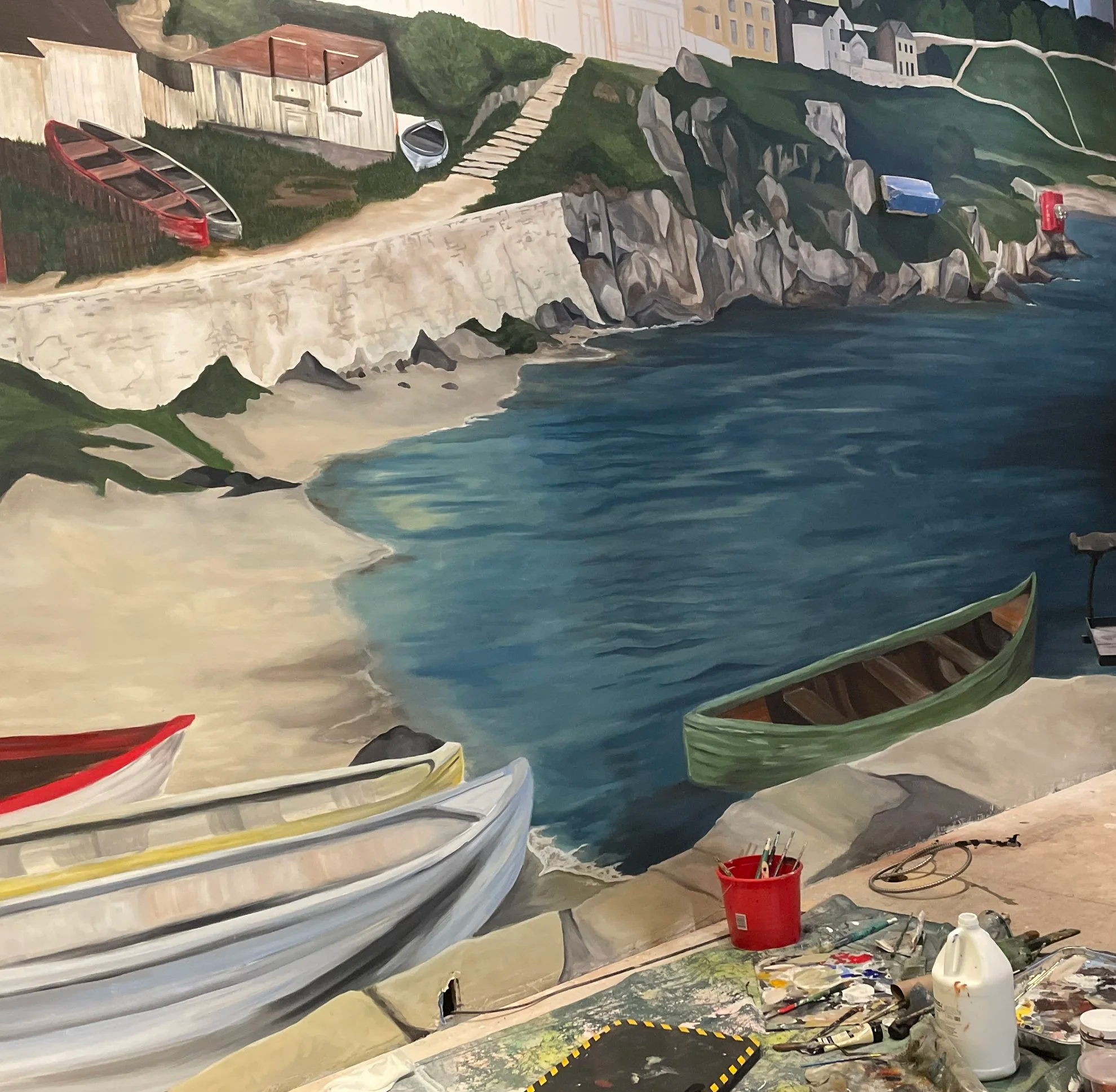 A painting of a coastal scene with boats on a rocky shore, a sandy beach, cliffs, green vegetation, and houses in the background. Artist supplies are visible in the foreground.