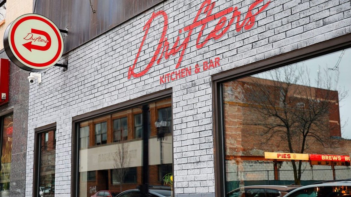 Exterior view of a restaurant called Lantons Kitchen & Bar, with white brick walls, large glass windows reflecting nearby buildings and trees, and a circular sign with a red arrow and the word 'Dutons' beside the restaurant sign.