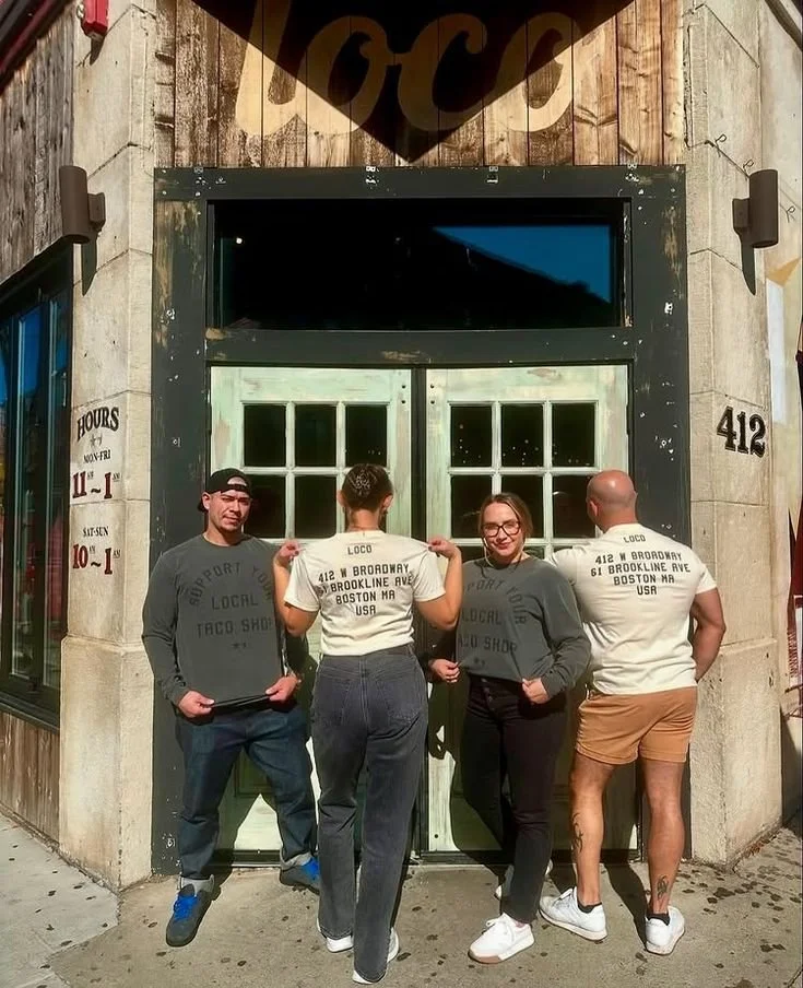 Four people standing in front of a restaurant with a sign above the door. Two are facing away, holding shirts with address and USA printed on the back. The other two face forward, smiling. The building has a dark door with window panes. The street ha