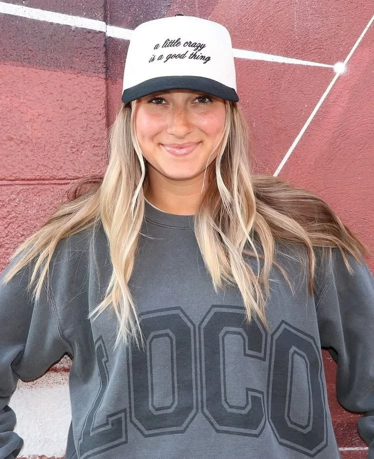 A young woman with long blonde hair wearing a gray sweatshirt with the word "LOCO" on it and a white trucker hat with black brim and text that reads "a little crazy is a good thing," custom merch designed for Loco Taqueria.