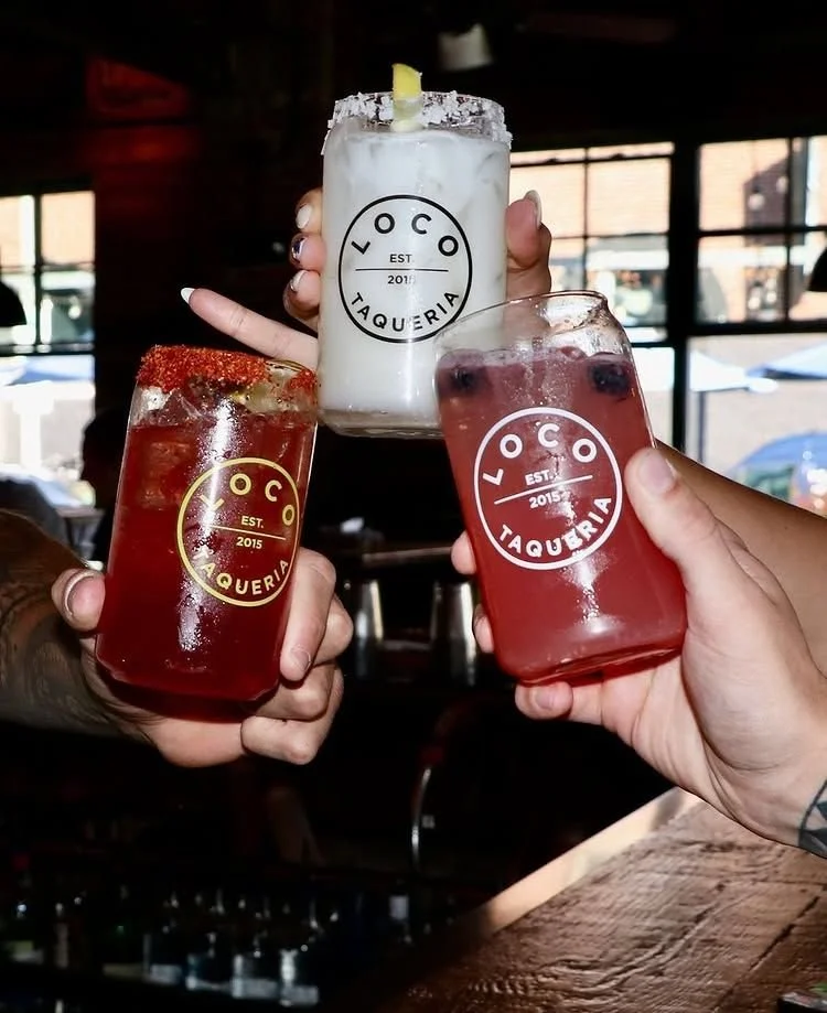 Three hands holding cocktails at a bar, two in red glasses with ice and garnishes and one in a tall white glass with a lemon wedge, all with 'LOCO TAQUERIA' logo.