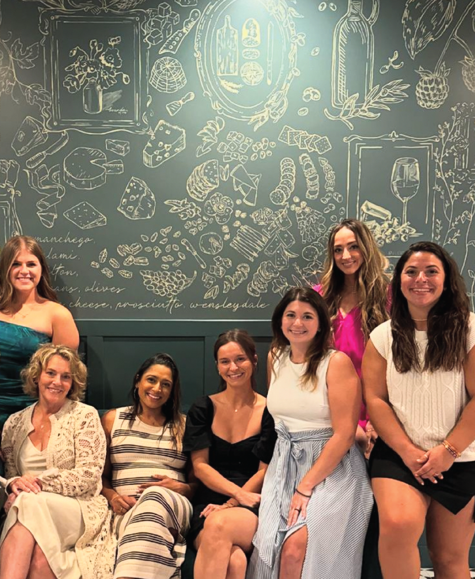 Group of six women smiling and posing for a photo in front of a food and wine themed mural in downtown Boston.