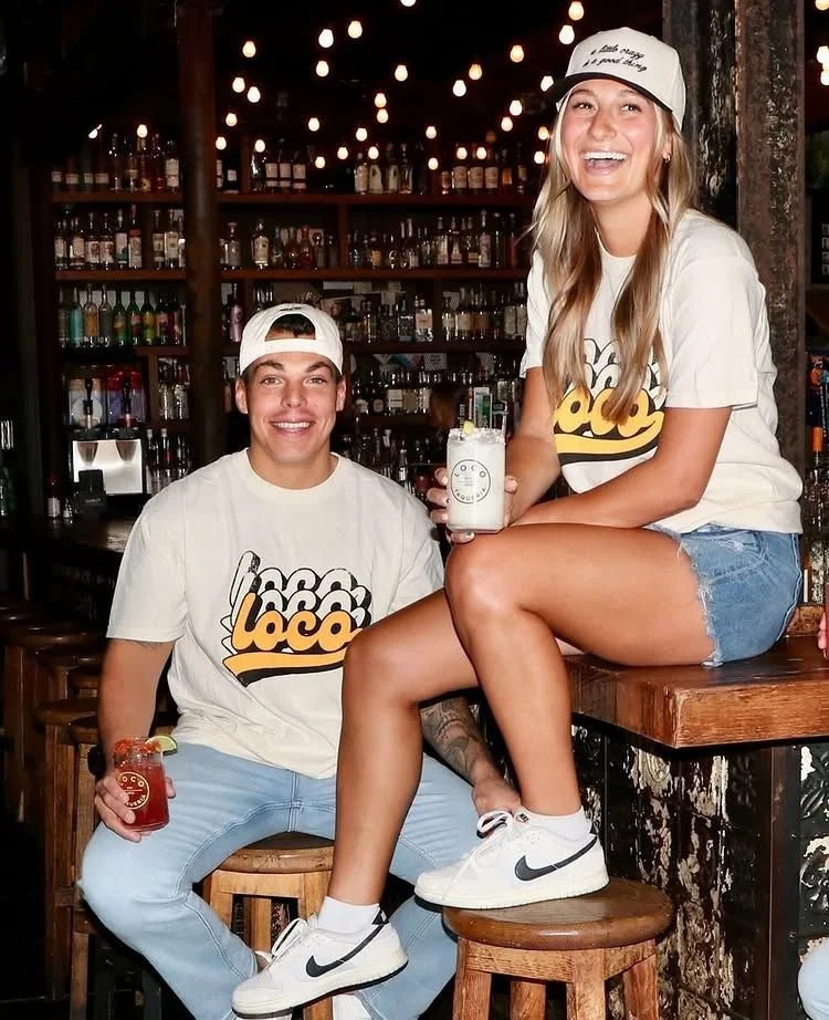 A smiling young woman sitting on a barstool with her legs crossed, holding a cocktail, and a smiling young man sitting on a stool beside her holding a cocktail, in a dimly lit bar with string lights and liquor bottles in the background.
