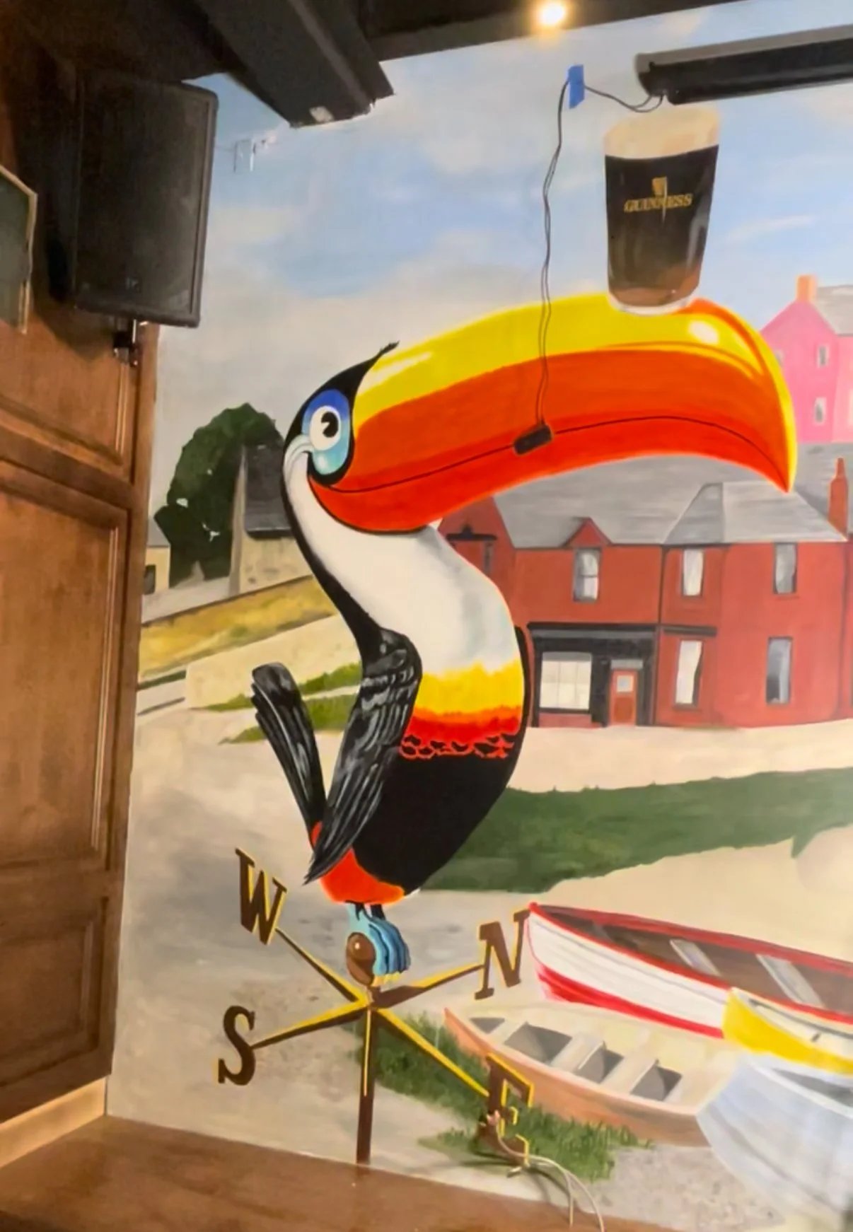Colorful wall mural of a toucan perched on a weather vane, with a pint of Guinness drink hanging from a wire above its beak, and a small boat on water nearby, with driftwood and houses in the background.