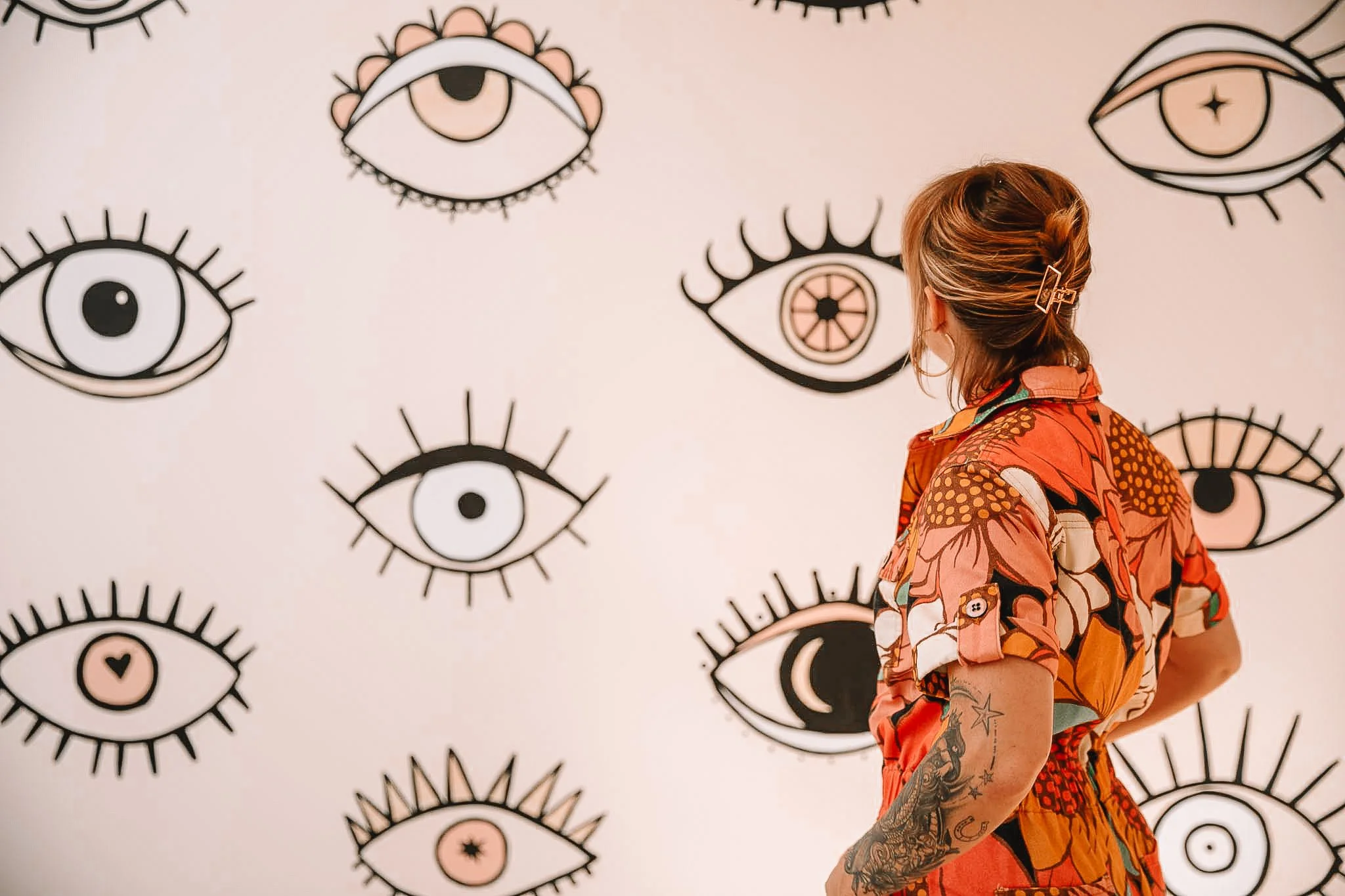 Person with tattoos on their arm looking at a wall with artistic eye illustrations in a gallery or exhibition.