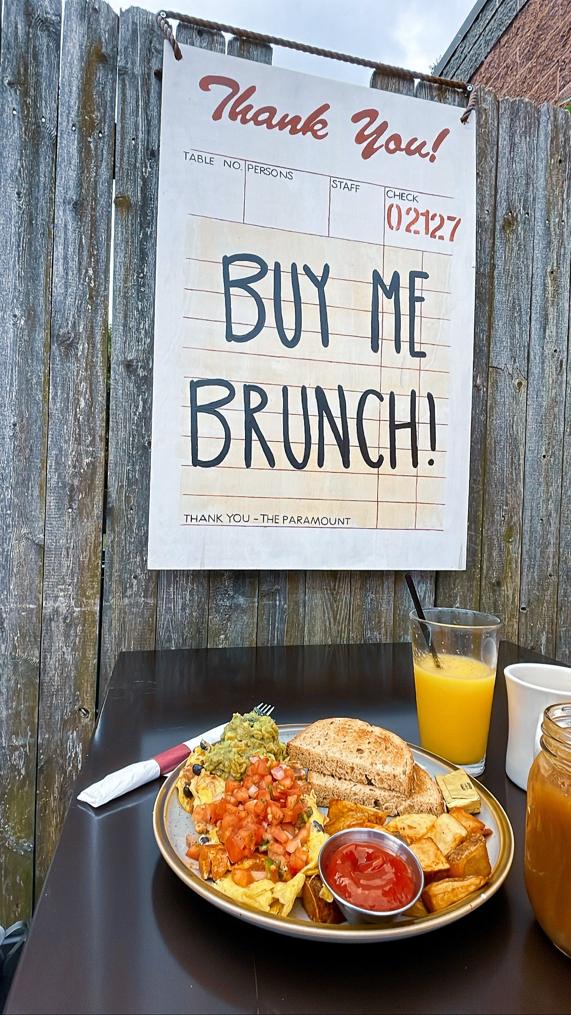 A plate of breakfast food including scrambled eggs topped with pico de gallo, guacamole, home fries with ketchup, a slice of toasted bread with butter, and drinks of orange juice and coffee. A sign in the background reads 'Thank You! Buy Me Brunch!' 