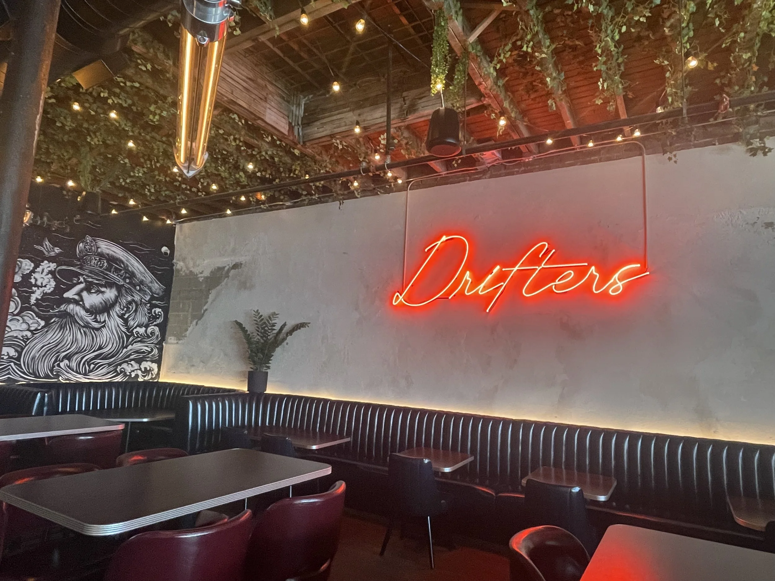 Interior of a modern bar or restaurant with a neon sign that says 'Drafters' on the wall, lit with string lights and decorated with greenery on the ceiling; there are tables and chairs along a black upholstered bench.