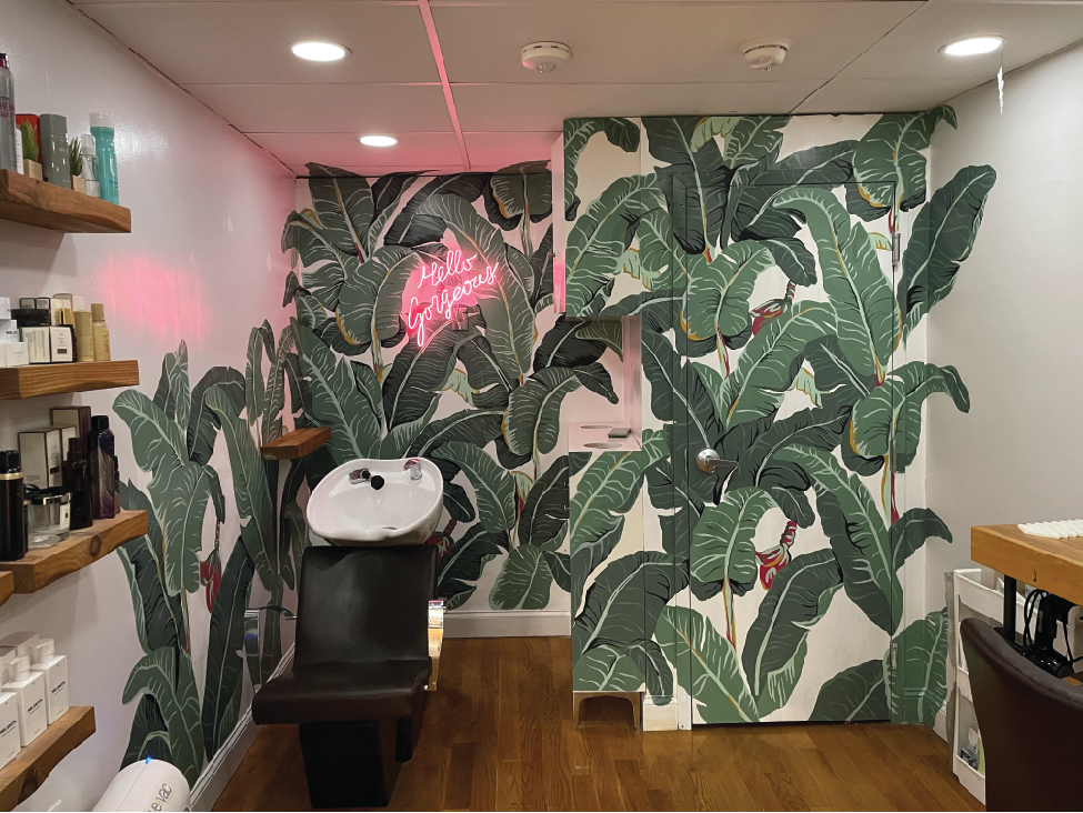 A salon with banana leaf mural wallpaper, a neon pink sign that says 'Hello Gorgeous', a white sink, a black salon chair, and wooden shelves with beauty products.