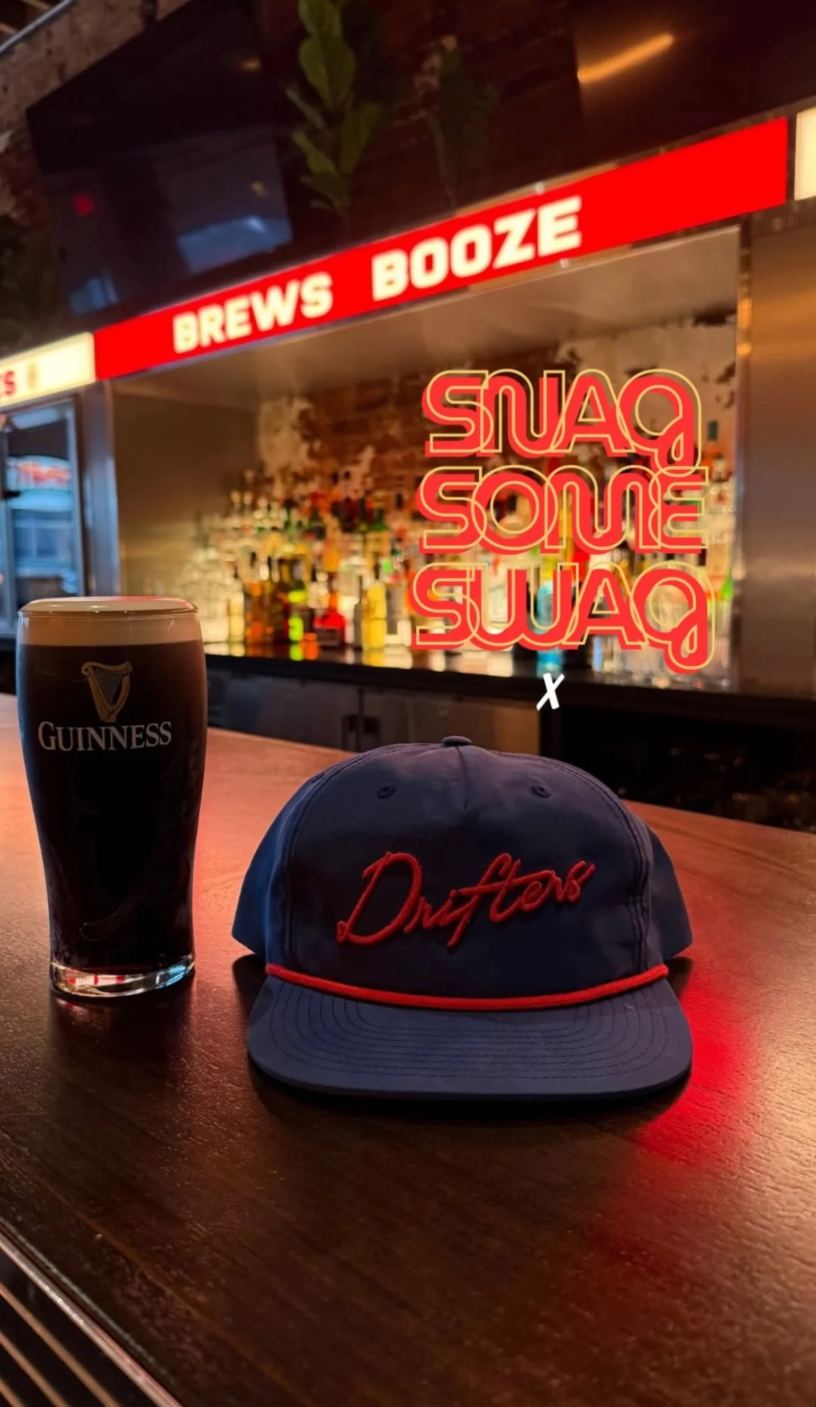 A pint of Guinness beer, a blue cap with red embroidered text, and a neon sign reading "SWAG SOME SWAG" on a bar counter with a backlit liquor shelf in the background. Merch and bar sign design by J Lenore Designs.