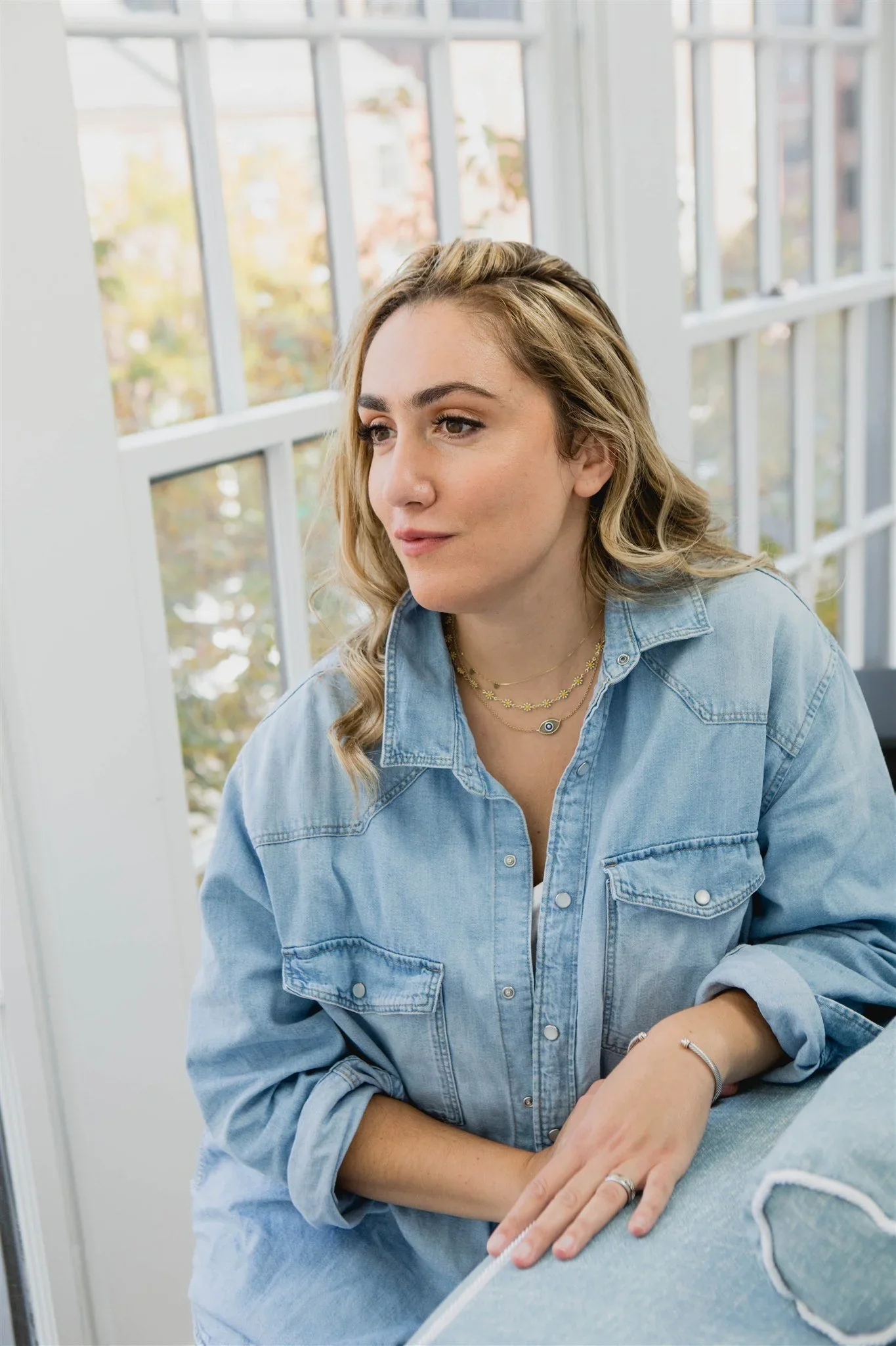 A young woman with blonde hair wearing a light blue denim shirt and layered necklaces sitting near a large window with white panes.