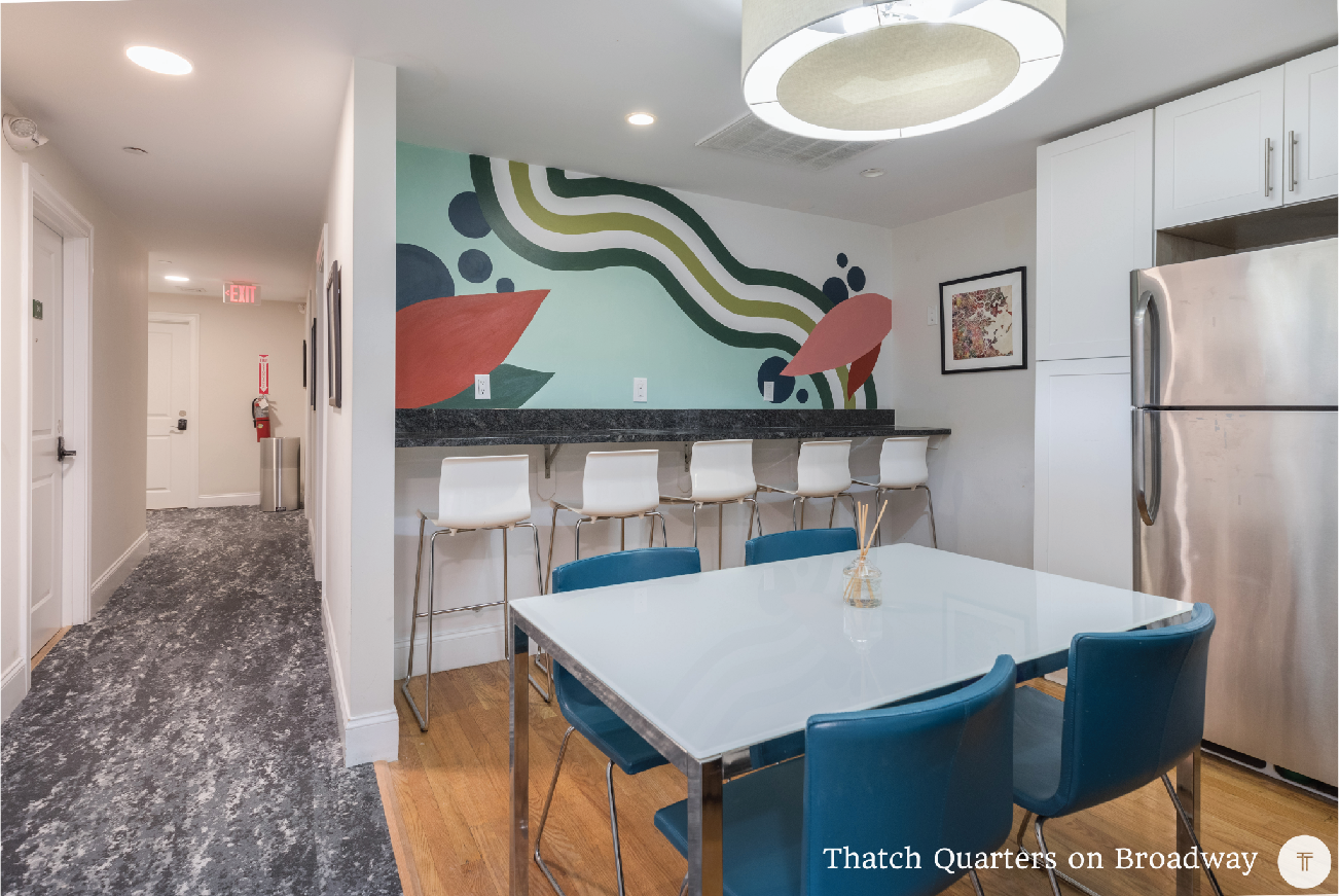 A modern dining area with a white table and four blue chairs, adjacent to a kitchen with white cabinets and a stainless steel refrigerator. There is a colorful abstract mural on the wall behind a bar counter with five white barstools, and a large cir