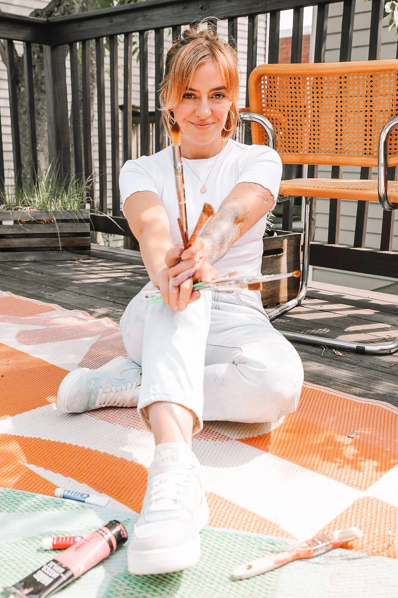 A woman sitting on an outdoor rug on a wooden deck, holding paintbrushes and smiling at the camera.