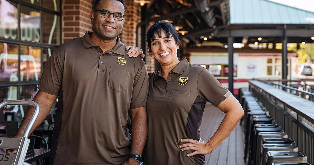 Two UPS employees, a man and a woman, standing outside a restaurant or cafe, smiling at the camera, wearing UPS uniforms.