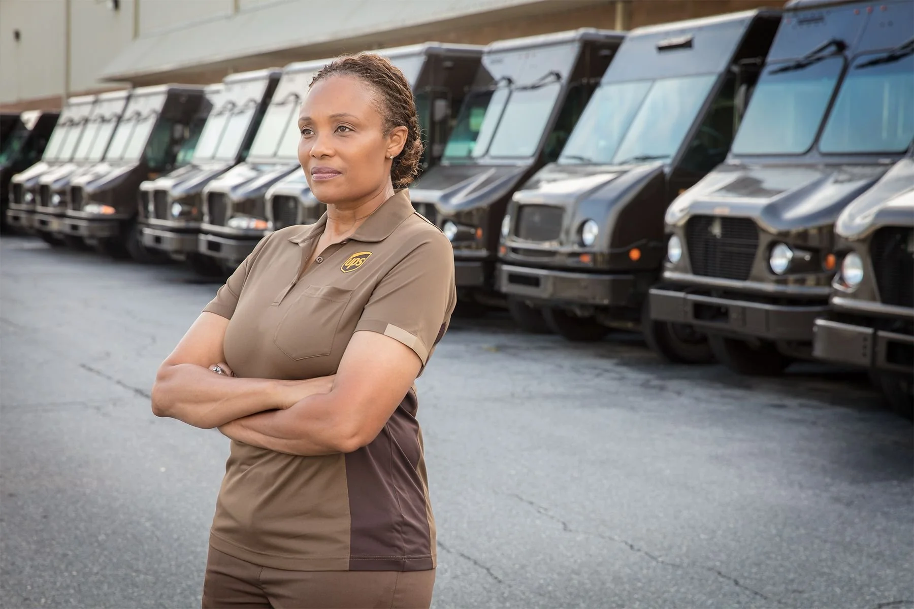 A woman in a UPS uniform standing with arms crossed in front of a row of delivery trucks.