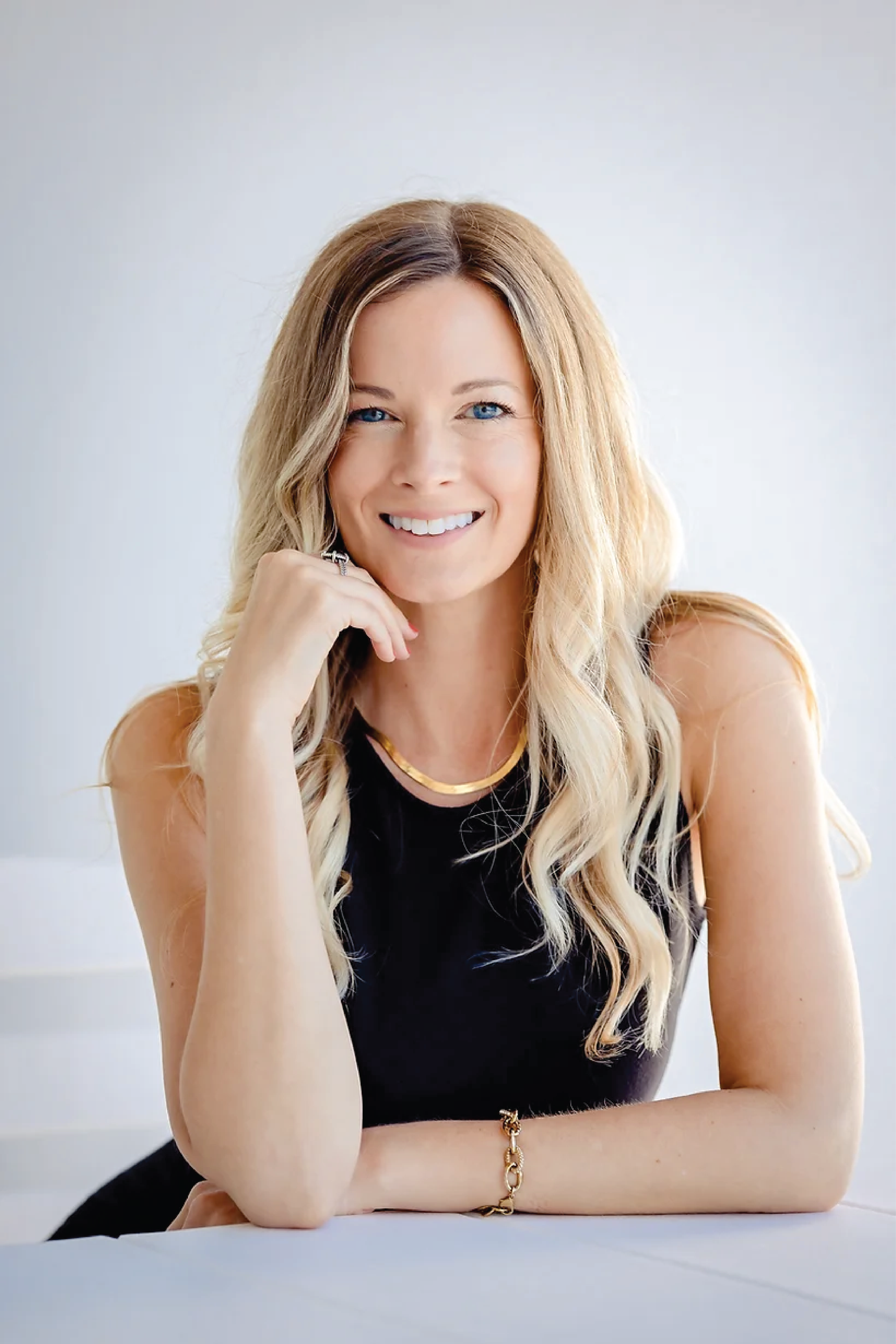 A woman with long blonde hair, blue eyes, and a bright smile is sitting at a white table, wearing a black sleeveless top, gold necklace, and bracelet, posing with her hand near her chin against a plain light-colored background.