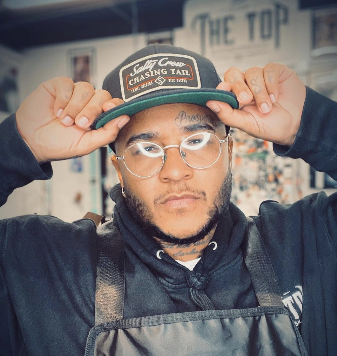 A young man with glasses adjusting his black cap, wearing a black hoodie and apron, in a tattoo studio.