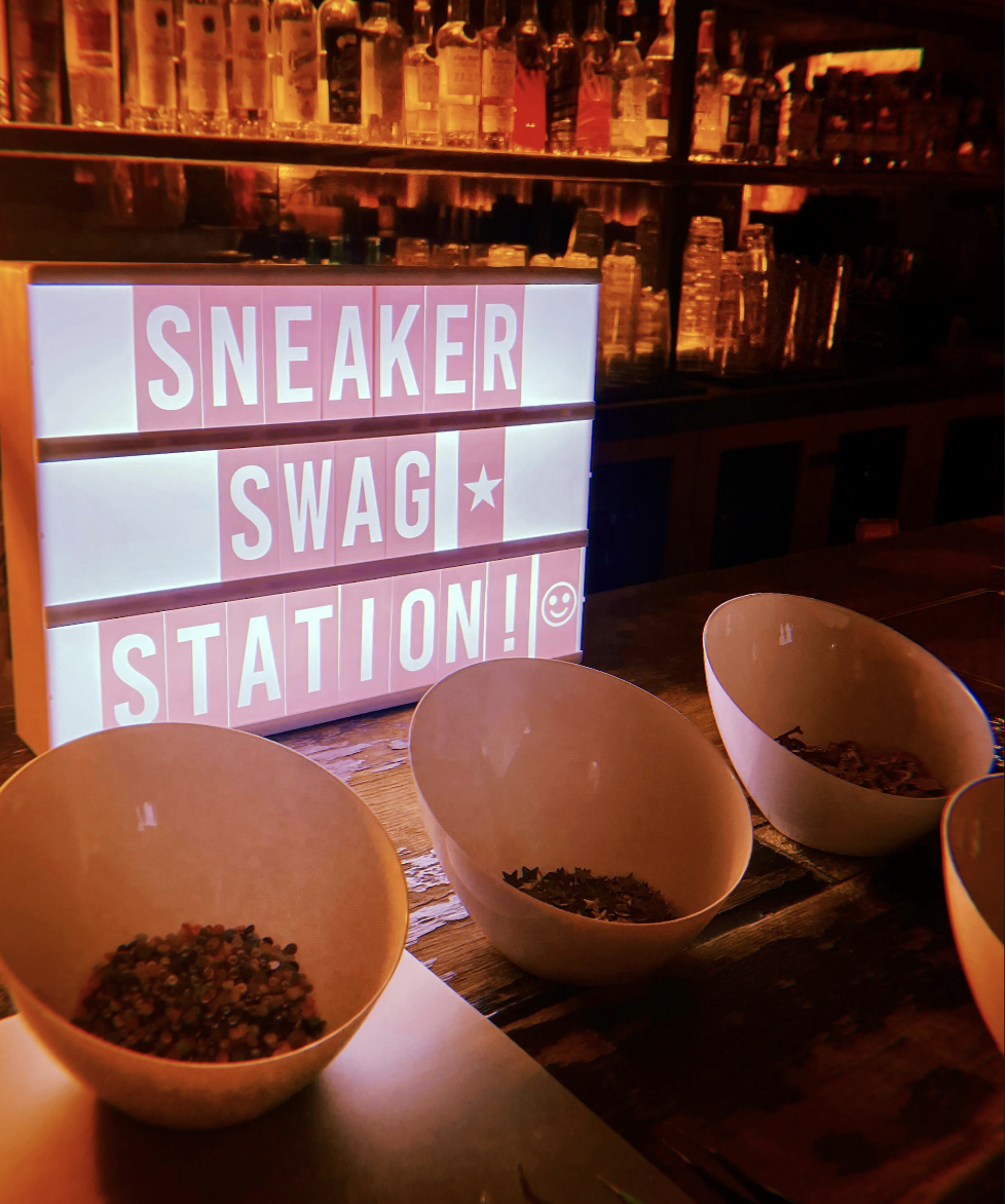 A light-up sign that reads 'SNEAKER SWAG STATION!' with a star and a smiley face emoji, part of a custom sneaker creative activation led by Jenna of J Lenore Designs.