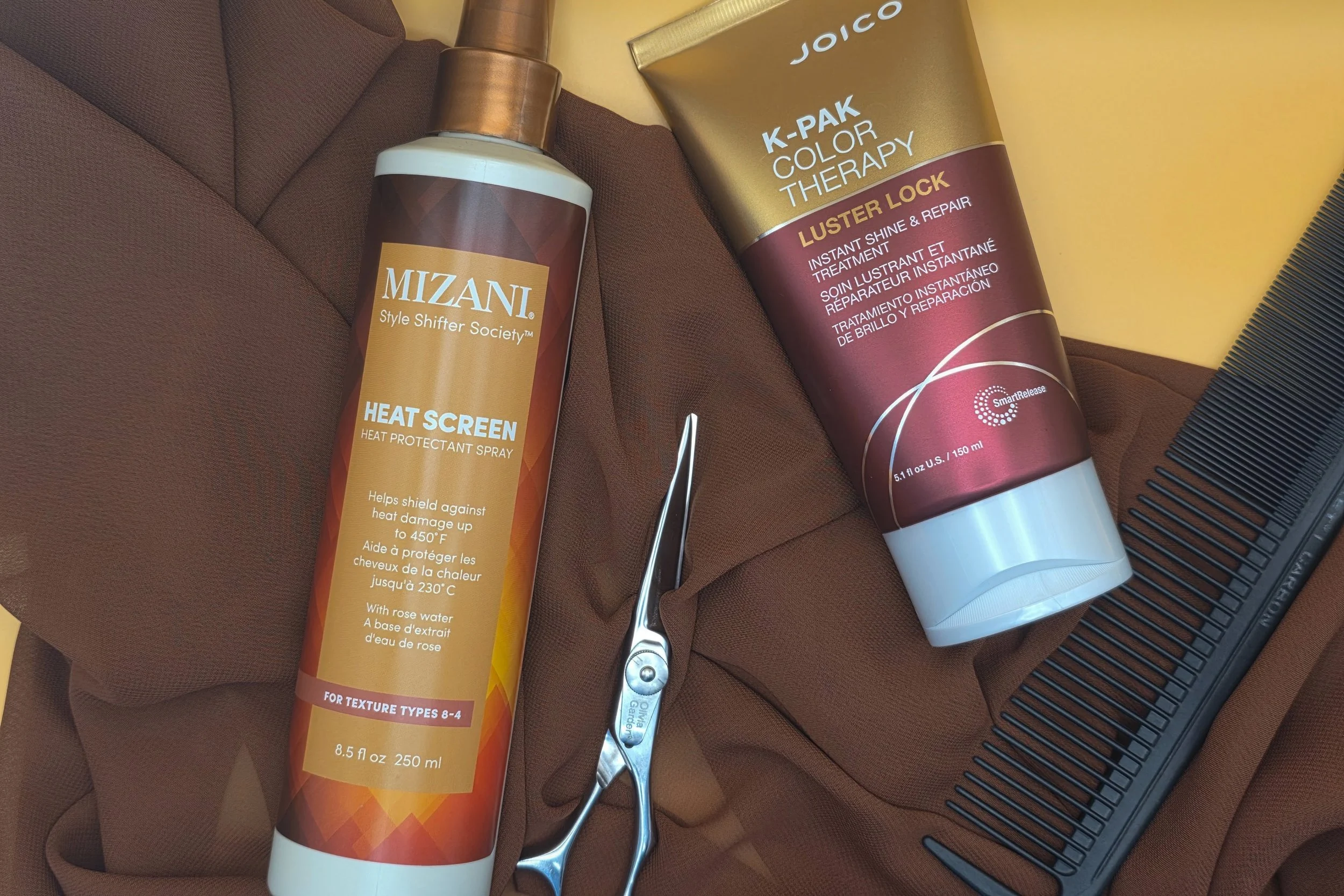 Hair styling tools and products including Mizani Heat Screen spray, Joico K-Pak Color Therapy Luster Lock treatment, a pair of hair cutting scissors, a black haircutting comb, and a brown cloak on a work surface.