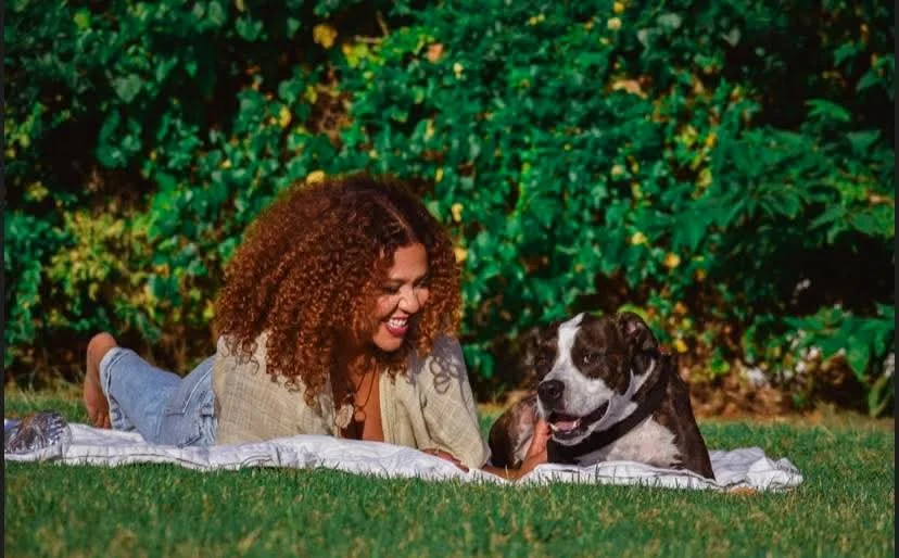 A woman with curly hair lying on a blanket on the grass, smiling and playing with a black and white dog in a lush green outdoor setting.