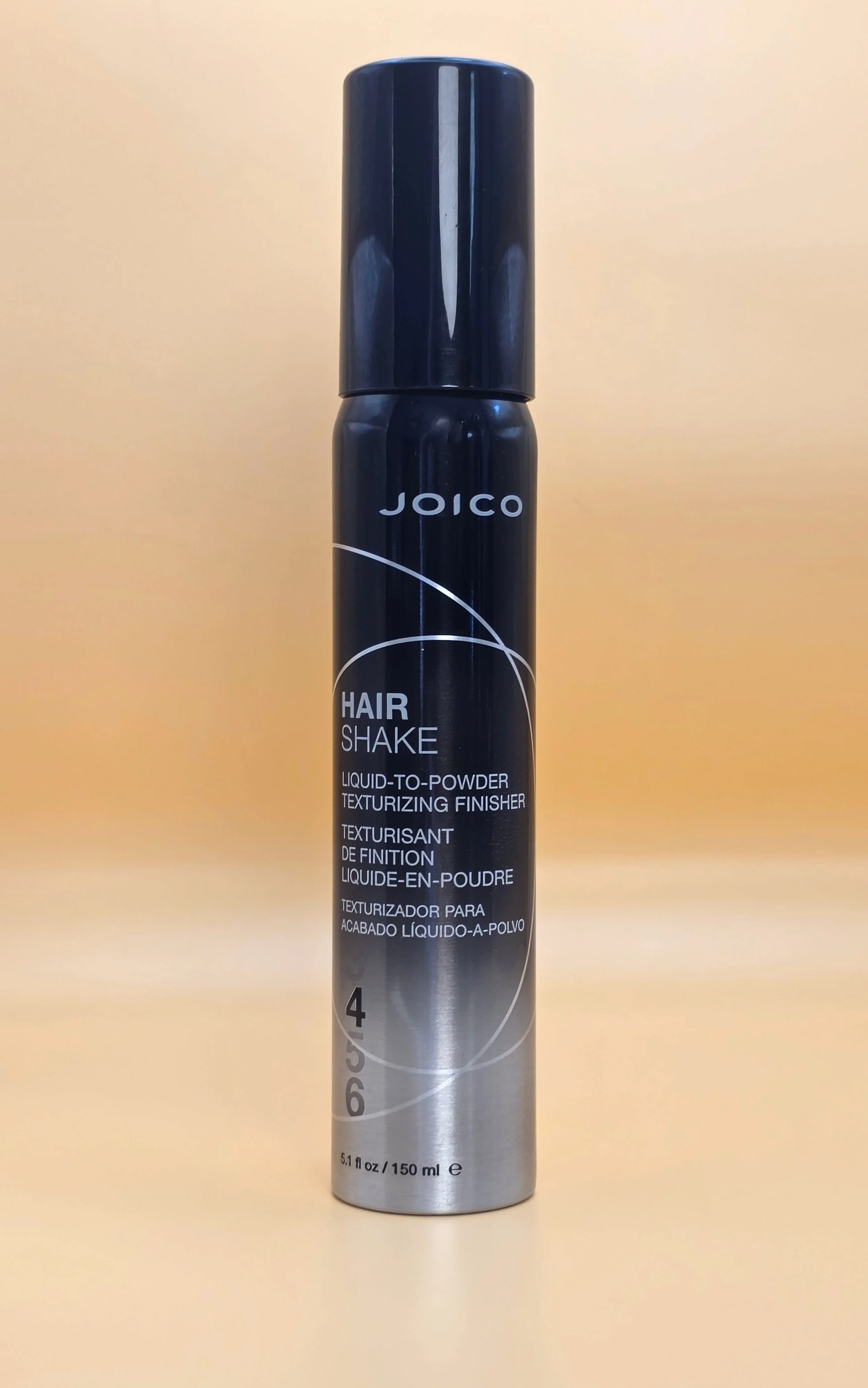 A can of Joico Hair Shake liquid-to-powder texturizing finisher with a black and silver design, placed on a light beige surface with a warm beige background.