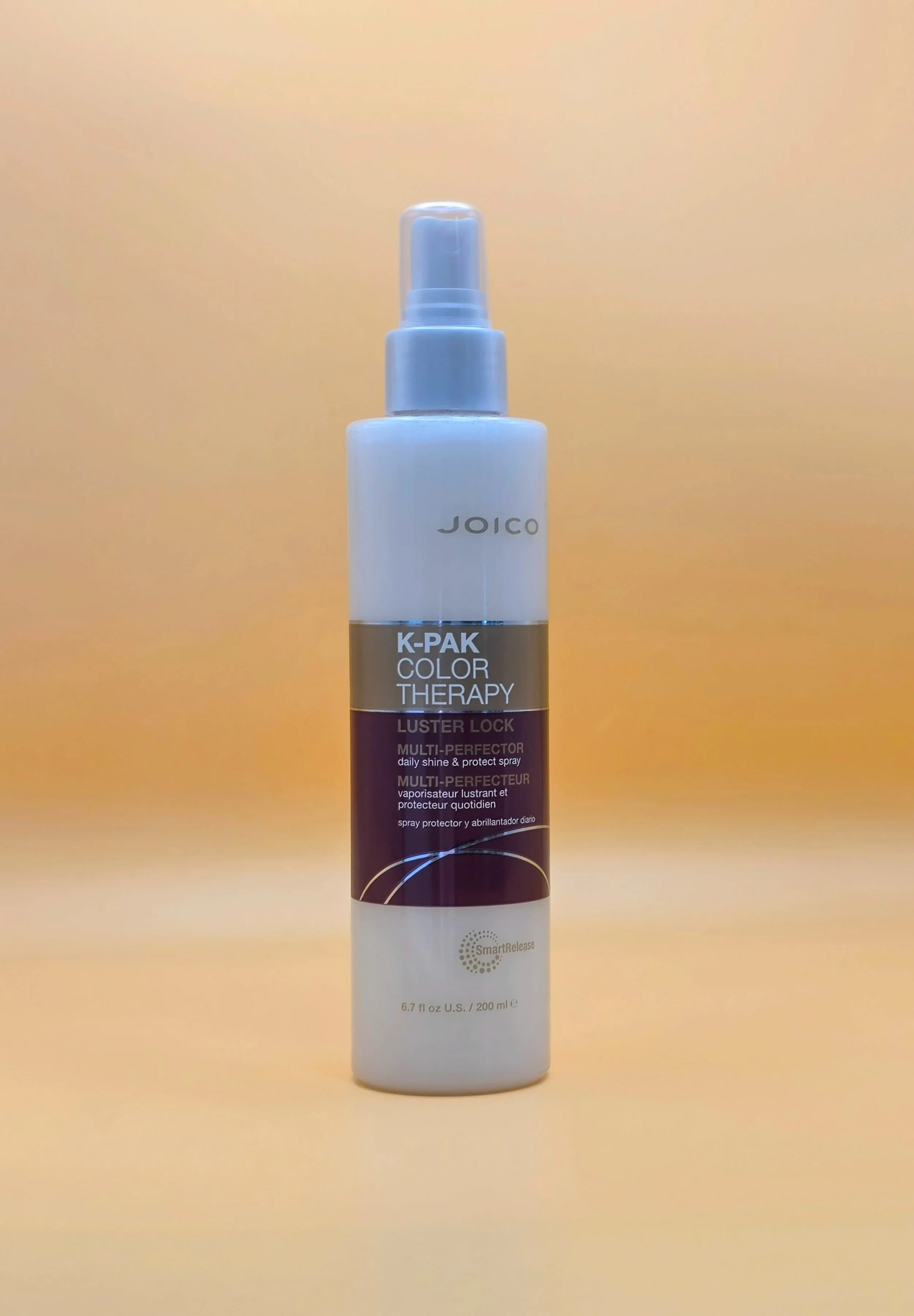 A white spray bottle labeled 'Joico K-Pak Color Therapy Luster Lock Multi-Perfection' with a silver band and a pump spray nozzle, standing on a yellow background.