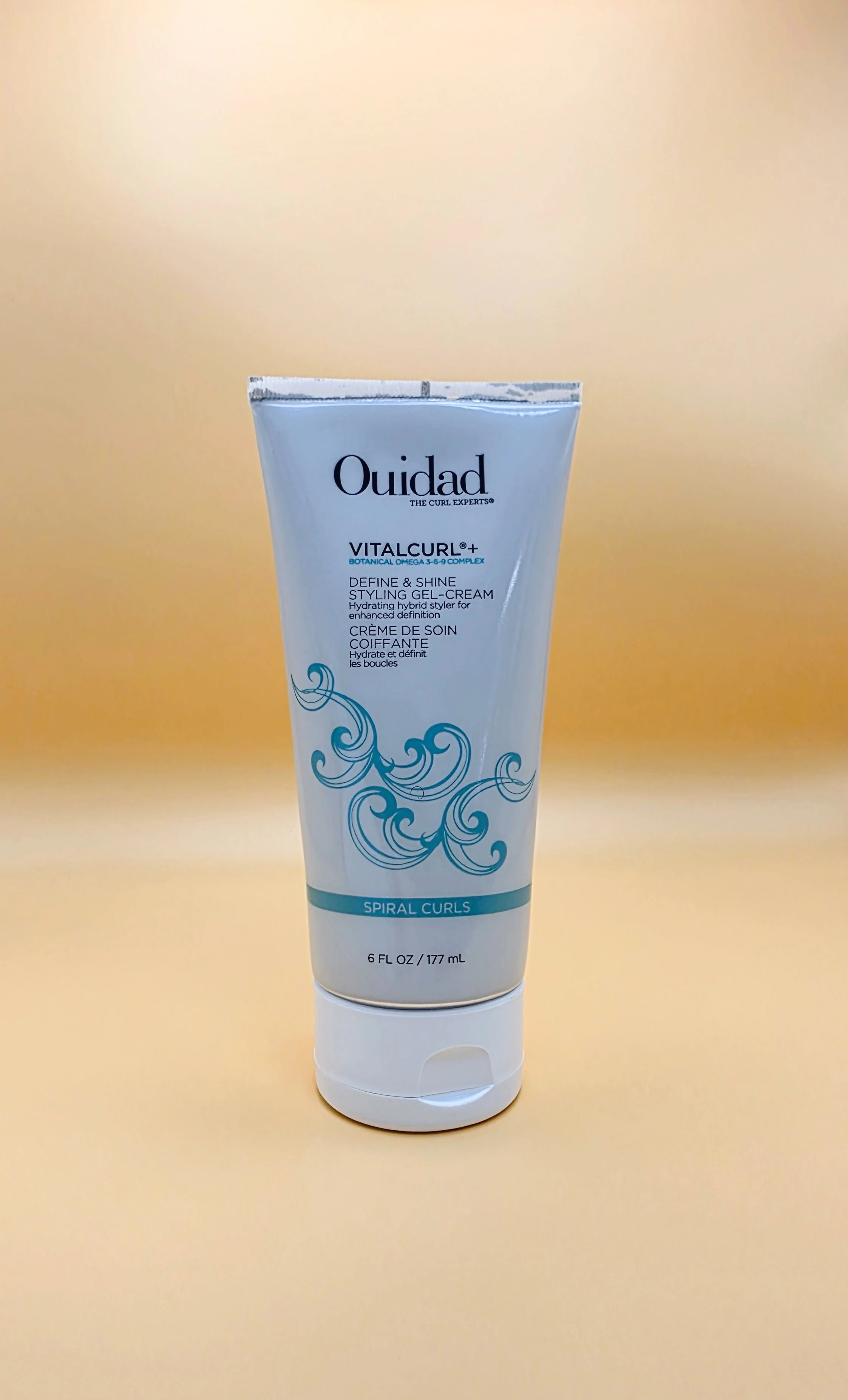 A white tube of Ouidad VitalCurl+ Define & Shine Styling Gel-Cream with blue swirl designs and text, standing upright on a light yellow background.