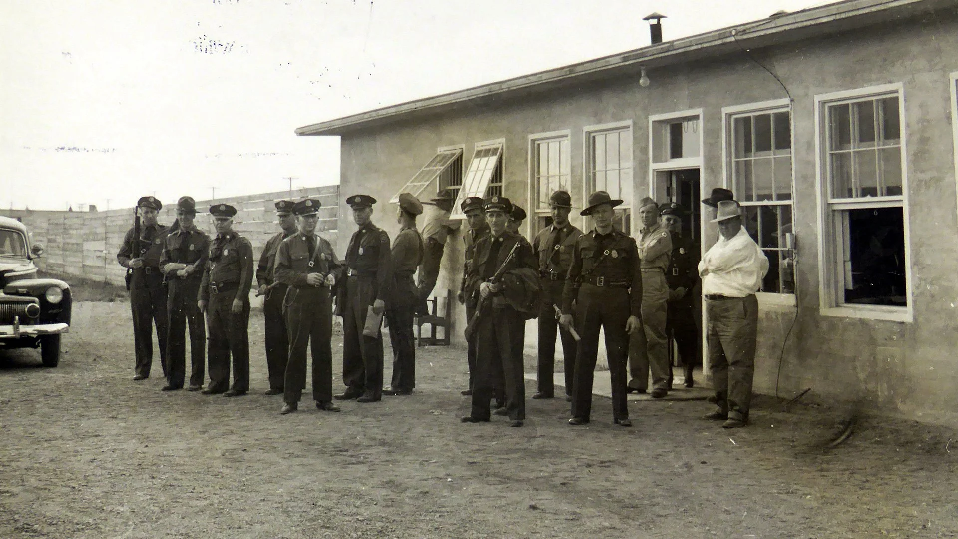 A black and white photo showing a group of police officers standing outside a building with open windows, next to a vintage car, in an outdoor setting.