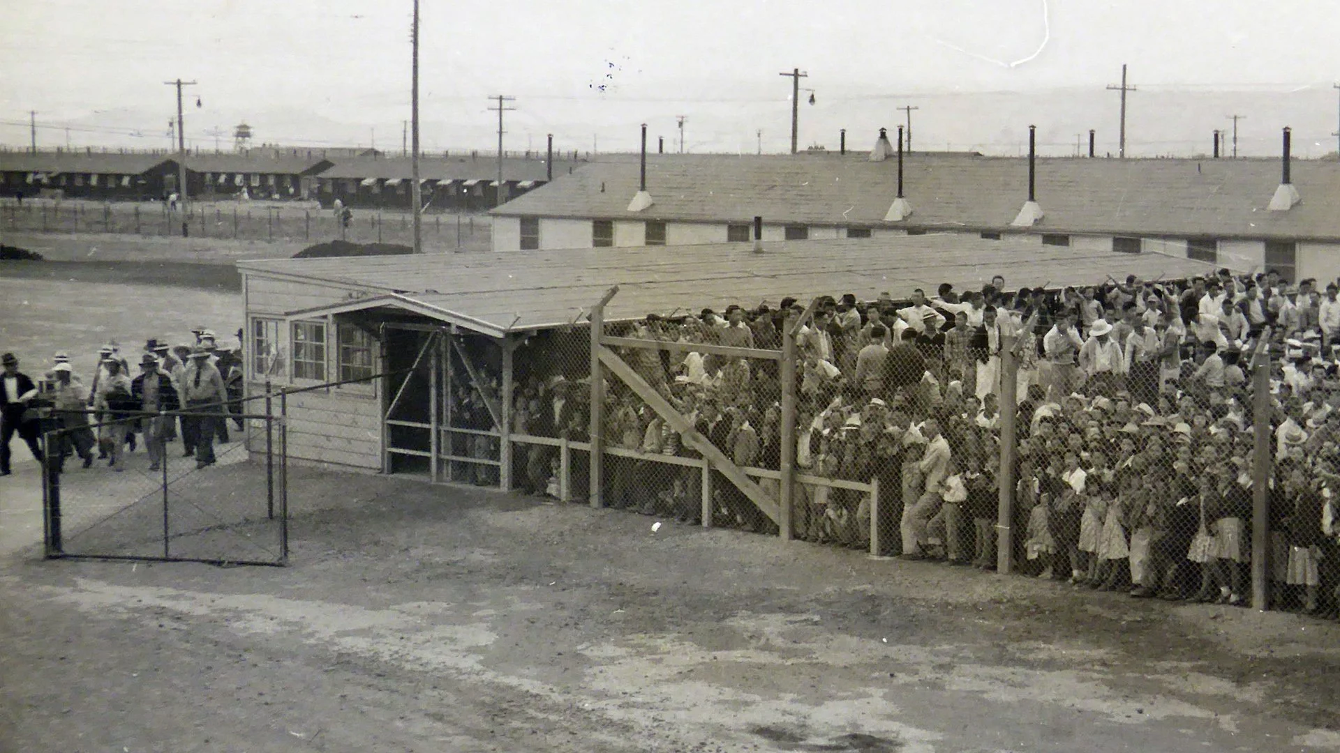 Black and white photo of a large crowd of people waiting behind a chain-link fence at a baseball game in a stadium, with some people sitting in the stands and others standing outside the fence.