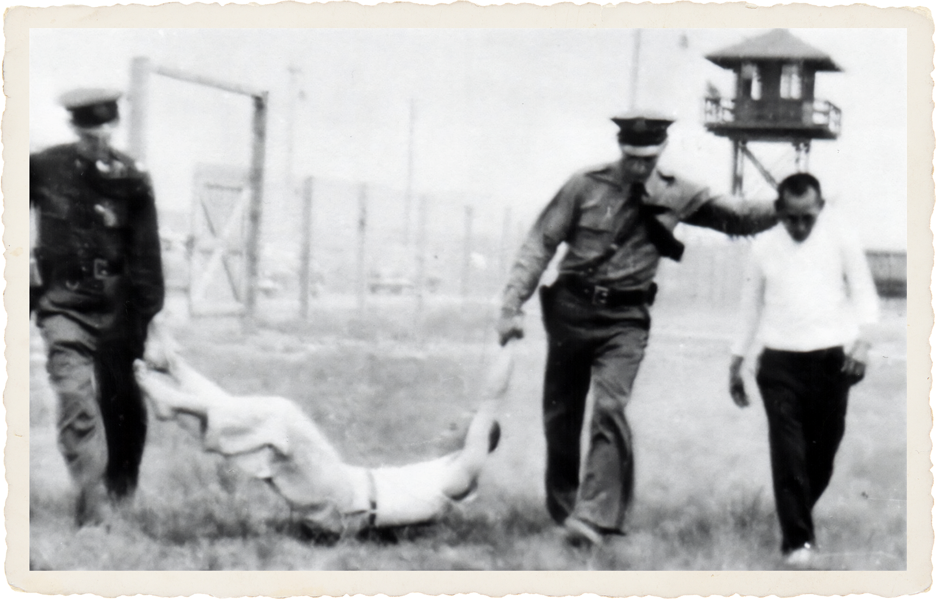 Black and white photo of two police officers and a woman. One officer is holding the woman's hair while the other officer appears to be detaining her. There is a guard tower in the background.