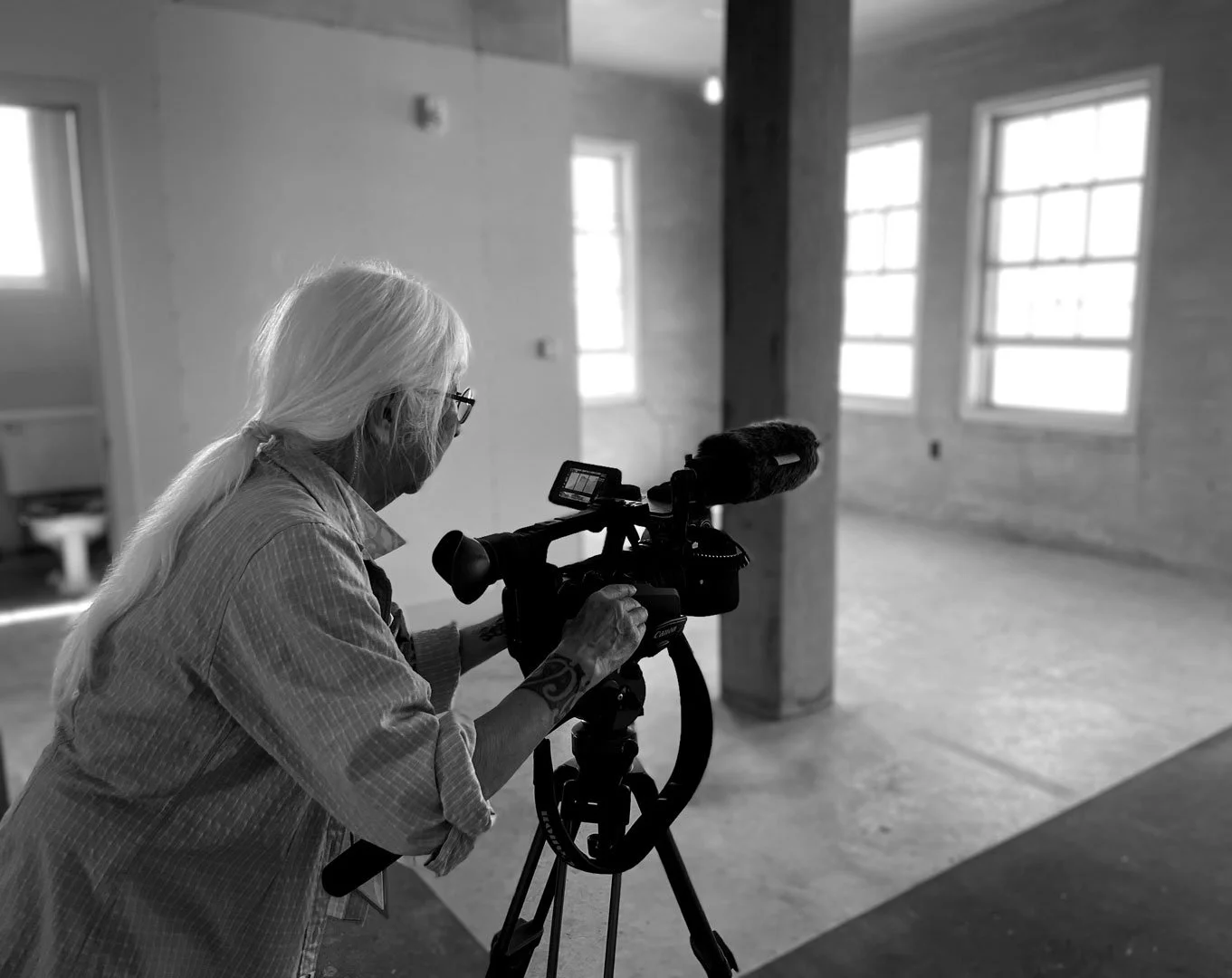 An elderly woman with long white hair wearing glasses and a striped shirt operates a professional video camera on a tripod inside a spacious, empty room with large windows and a wooden beam.