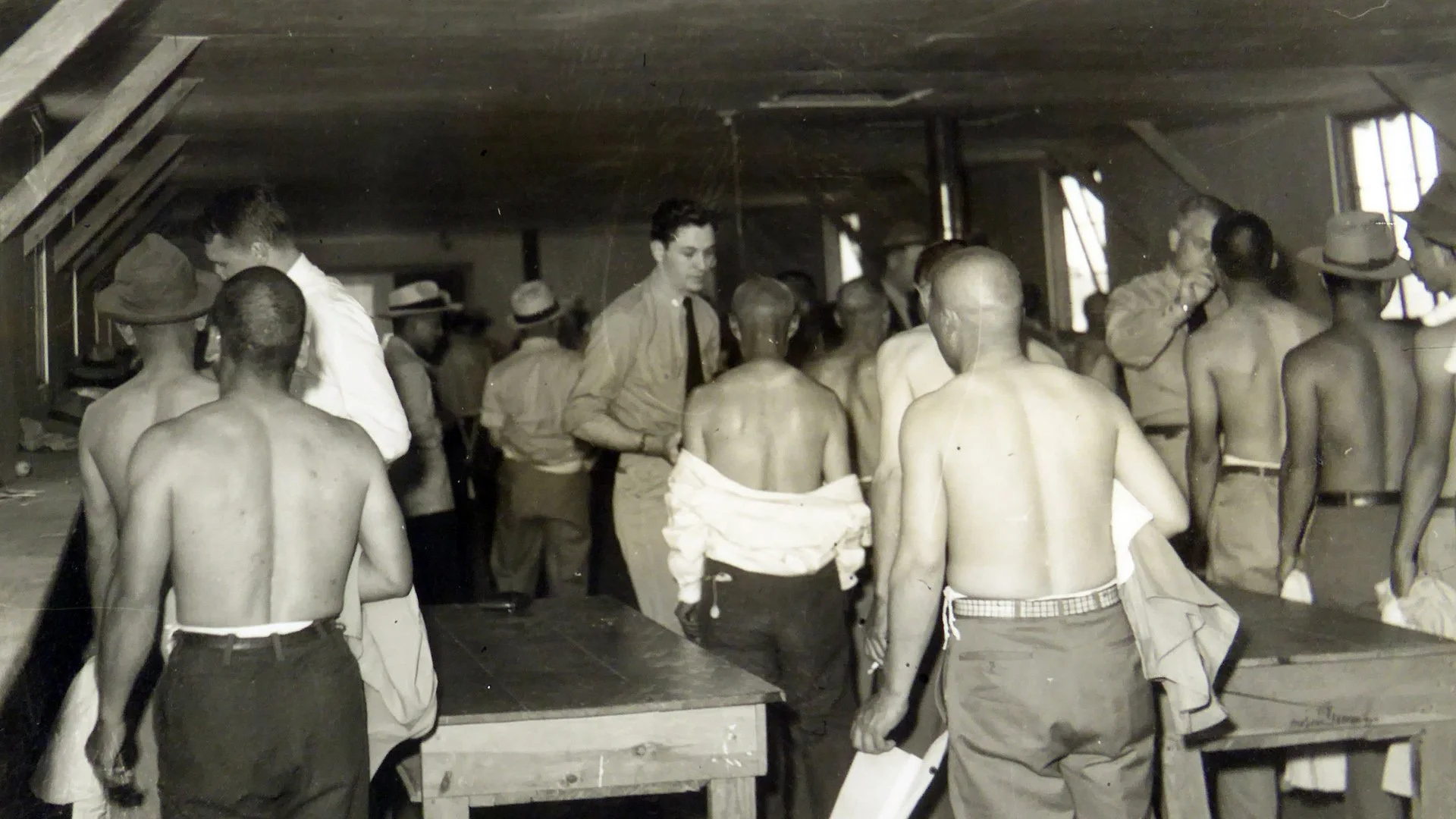 Black and white photograph of a scene inside a military barracks, showing shirtless soldiers with shaved heads, and officers in uniform conducting registration or administrative work.