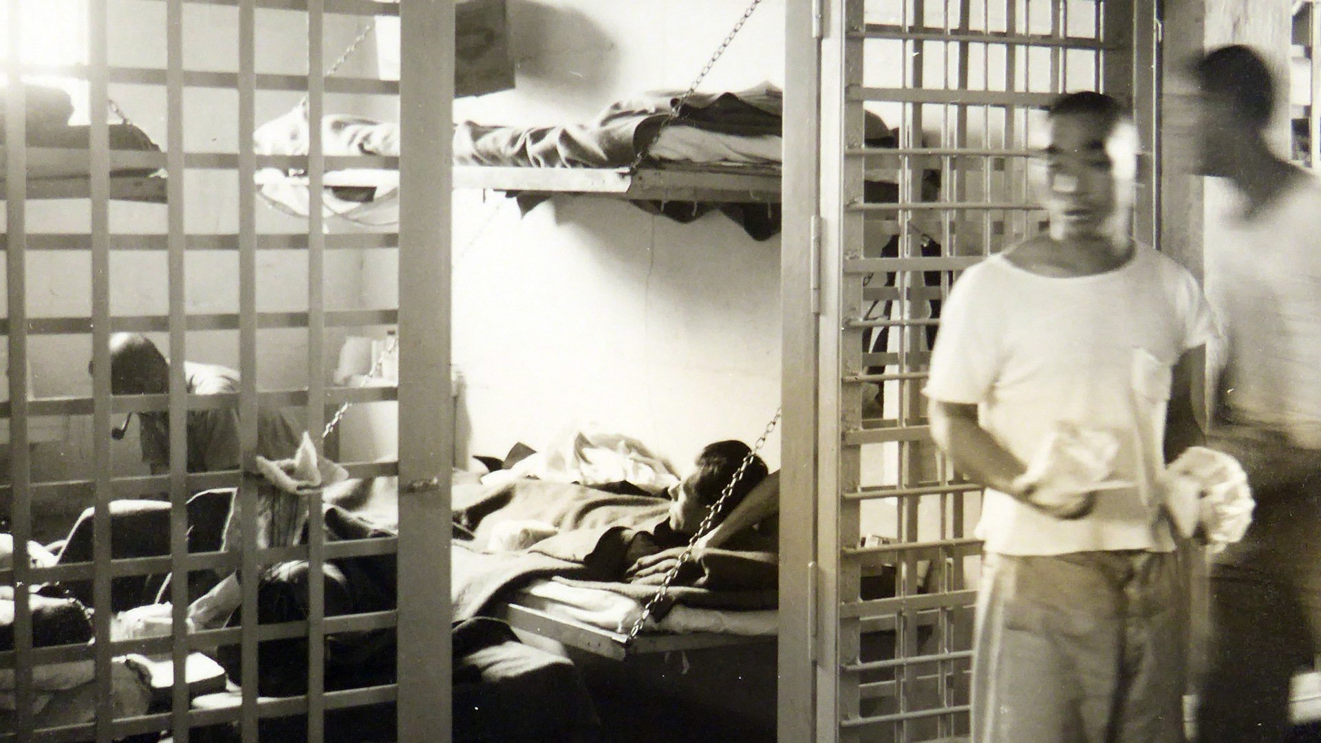 People inside a prison cell with metal bars, some lying on beds and one sitting up, in a dimly lit prison cell. Two individuals are walking outside the cell.