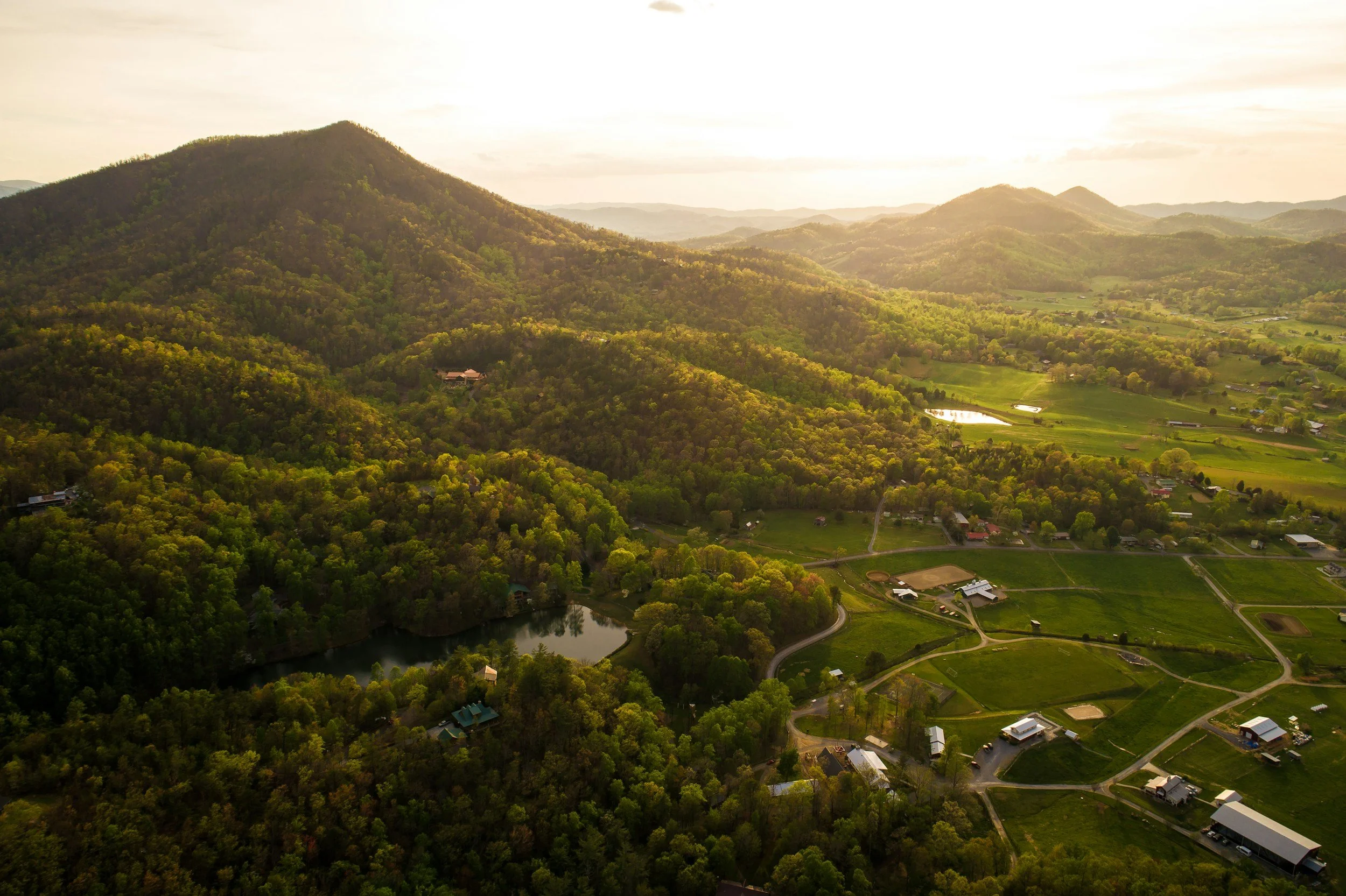 Aerial view of a rural landscape with green hills, a lake, open fields, and farm buildings during sunset.