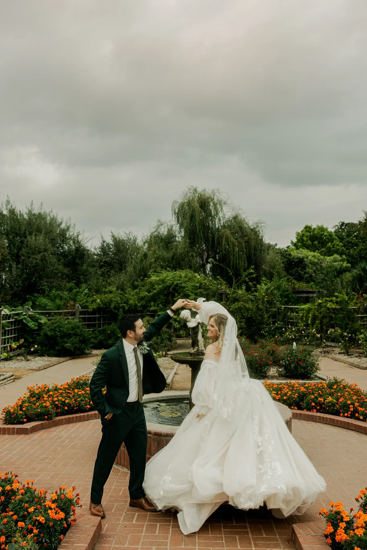 A bride and groom dancing outdoors in a garden with a fountain, surrounded by flowers and greenery.