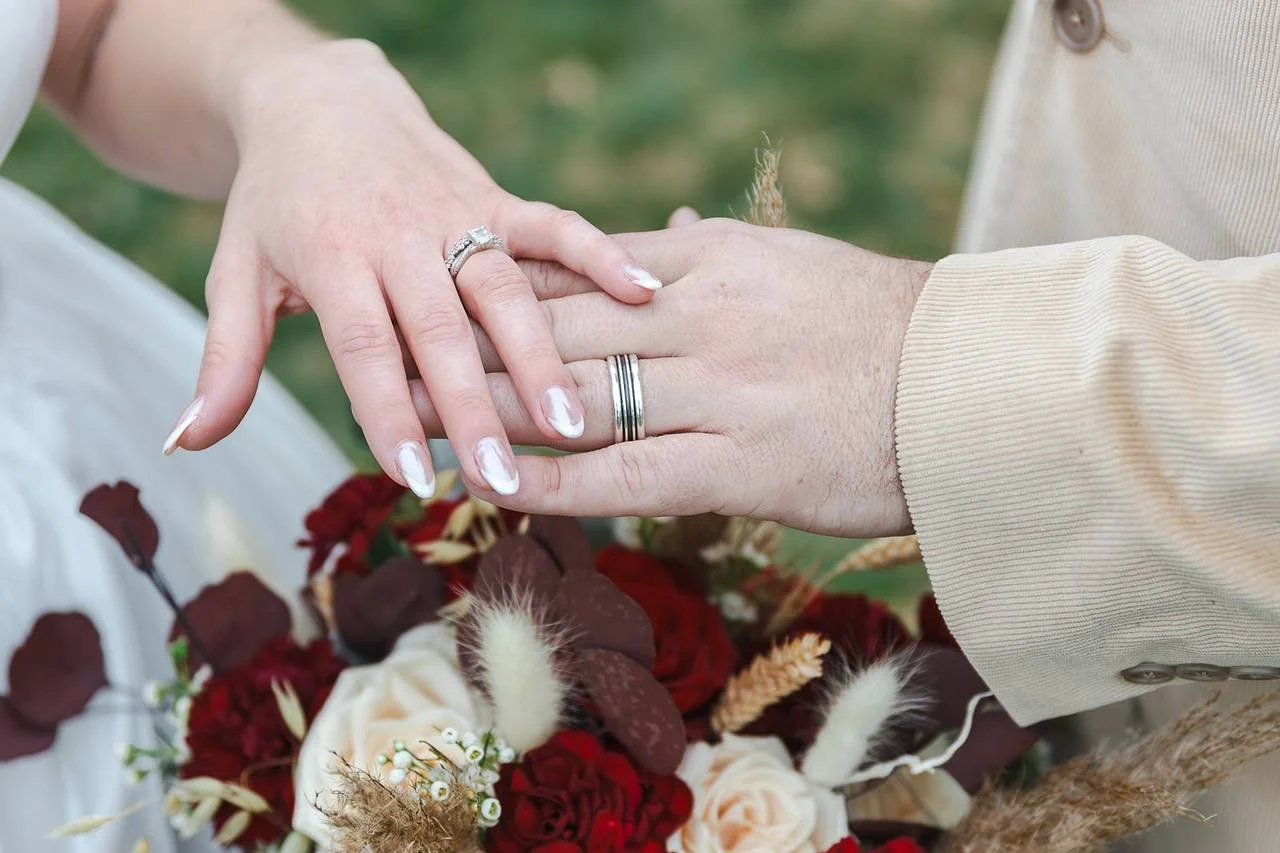 Close-up of a couple holding hands with wedding rings, a floral bouquet with red and white roses, and some dried flowers in the background.