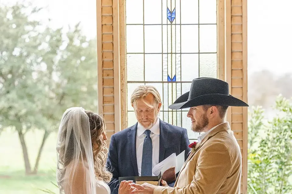 A wedding ceremony taking place indoors near a stained glass window with two people holding hands and a man in a suit and hat officiating.