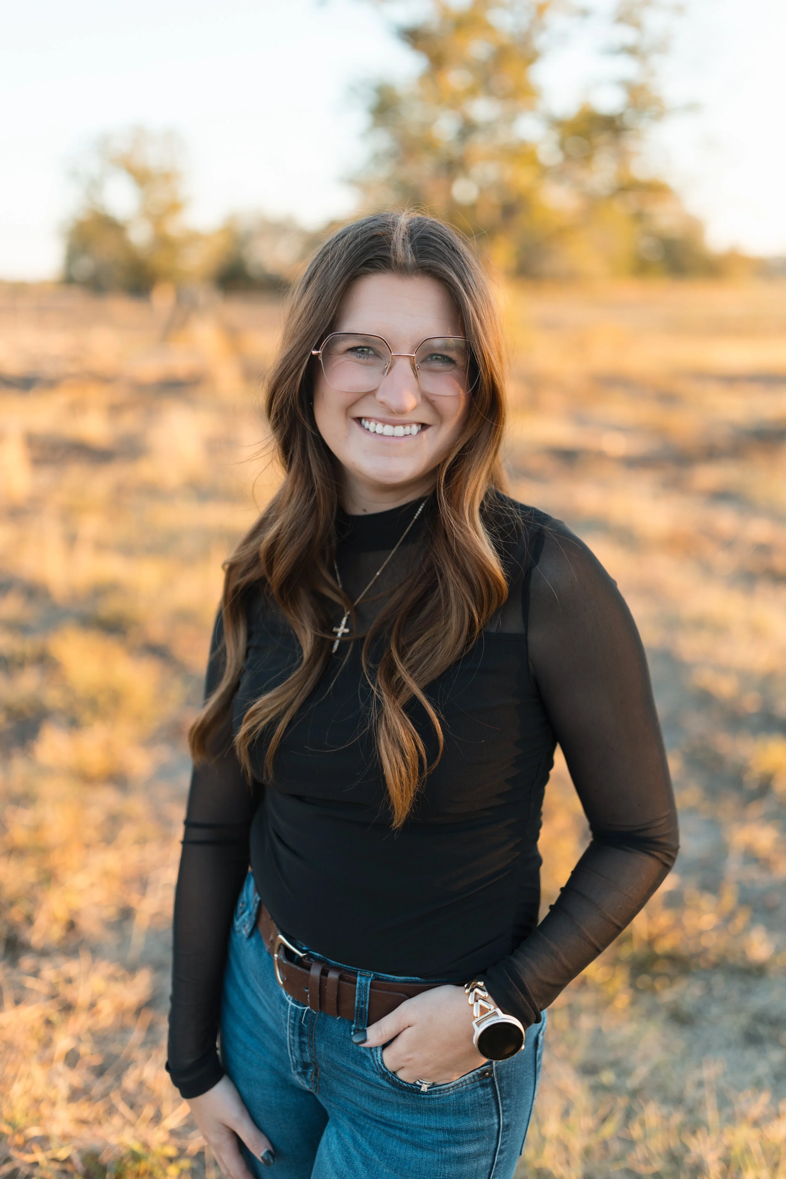 Smiling woman with long wavy brown hair wearing glasses, a black top, blue jeans, and a wristwatch, standing outdoors at sunset with trees in the background.