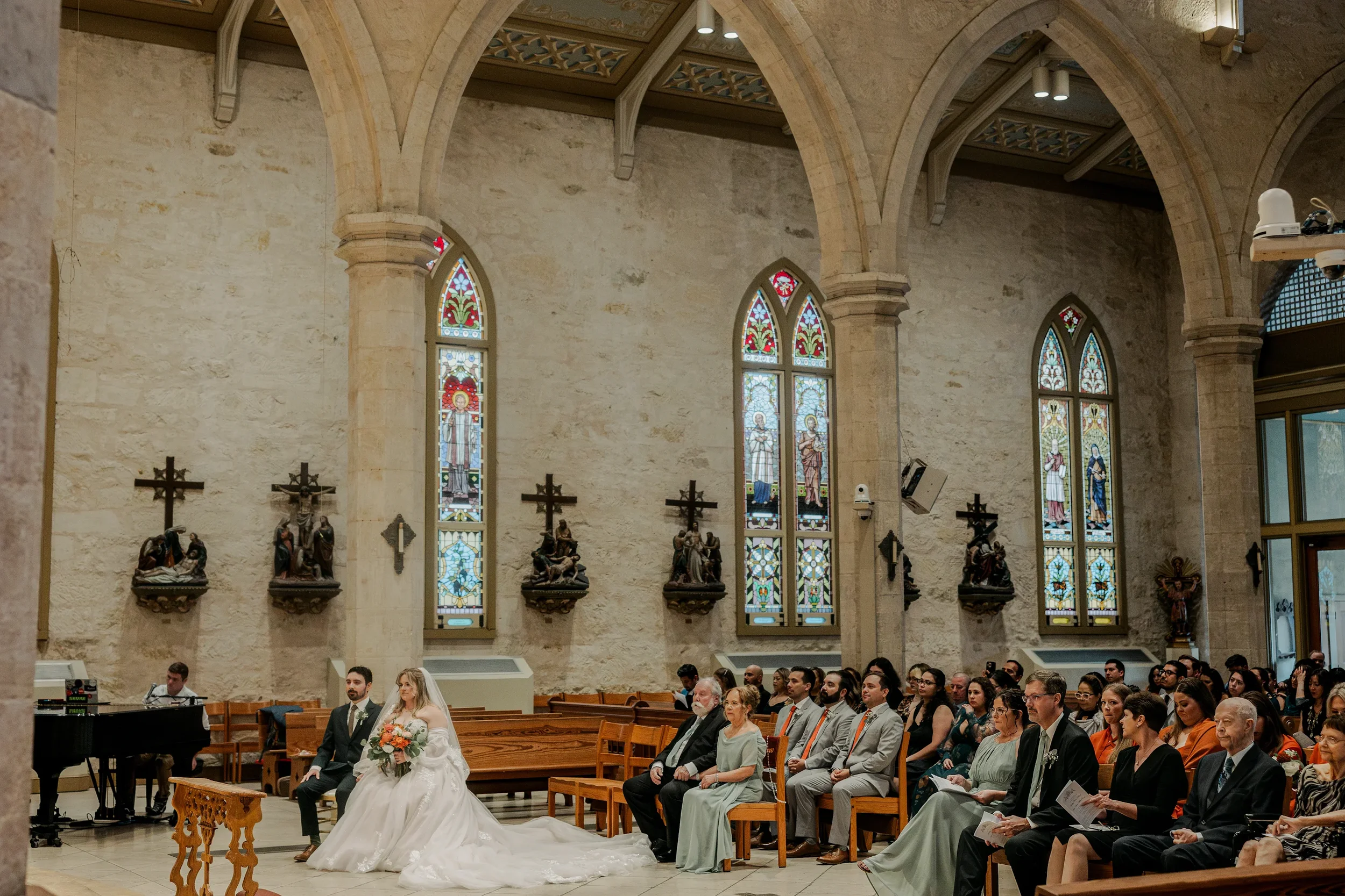 A wedding ceremony inside a church with stained glass windows, featuring a bride in a white wedding gown holding a bouquet, a groom in a dark suit, and guests seated in wooden pews, some wearing formal attire.