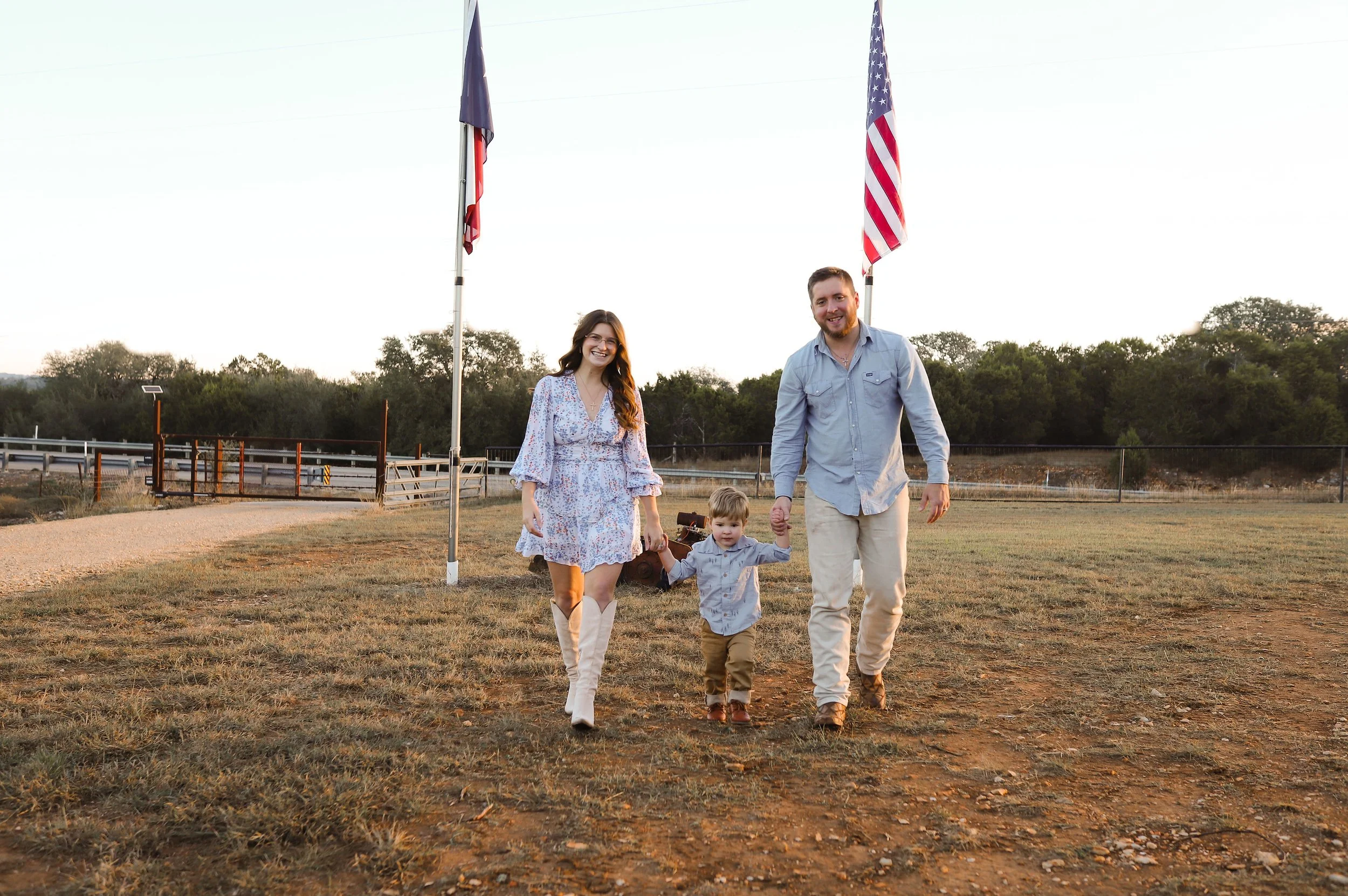A family of three walking hand-in-hand on a dirt path outdoors at sunset, with American and Texas flags in the background.