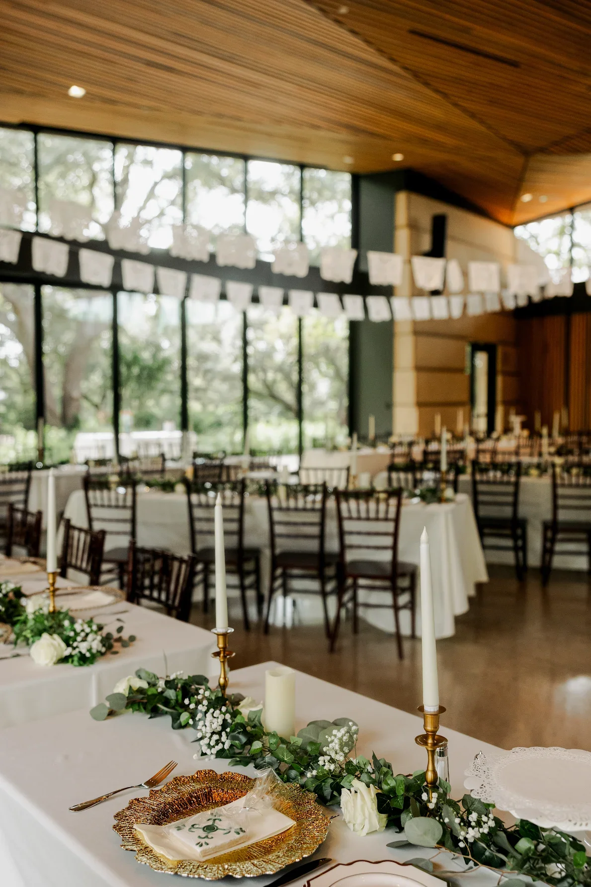 Elegant event space decorated with white tablecloths, greenery, white flowers, tall white candles, and gold candlesticks, set for a celebration, with a modern wooden ceiling and large windows in the background.
