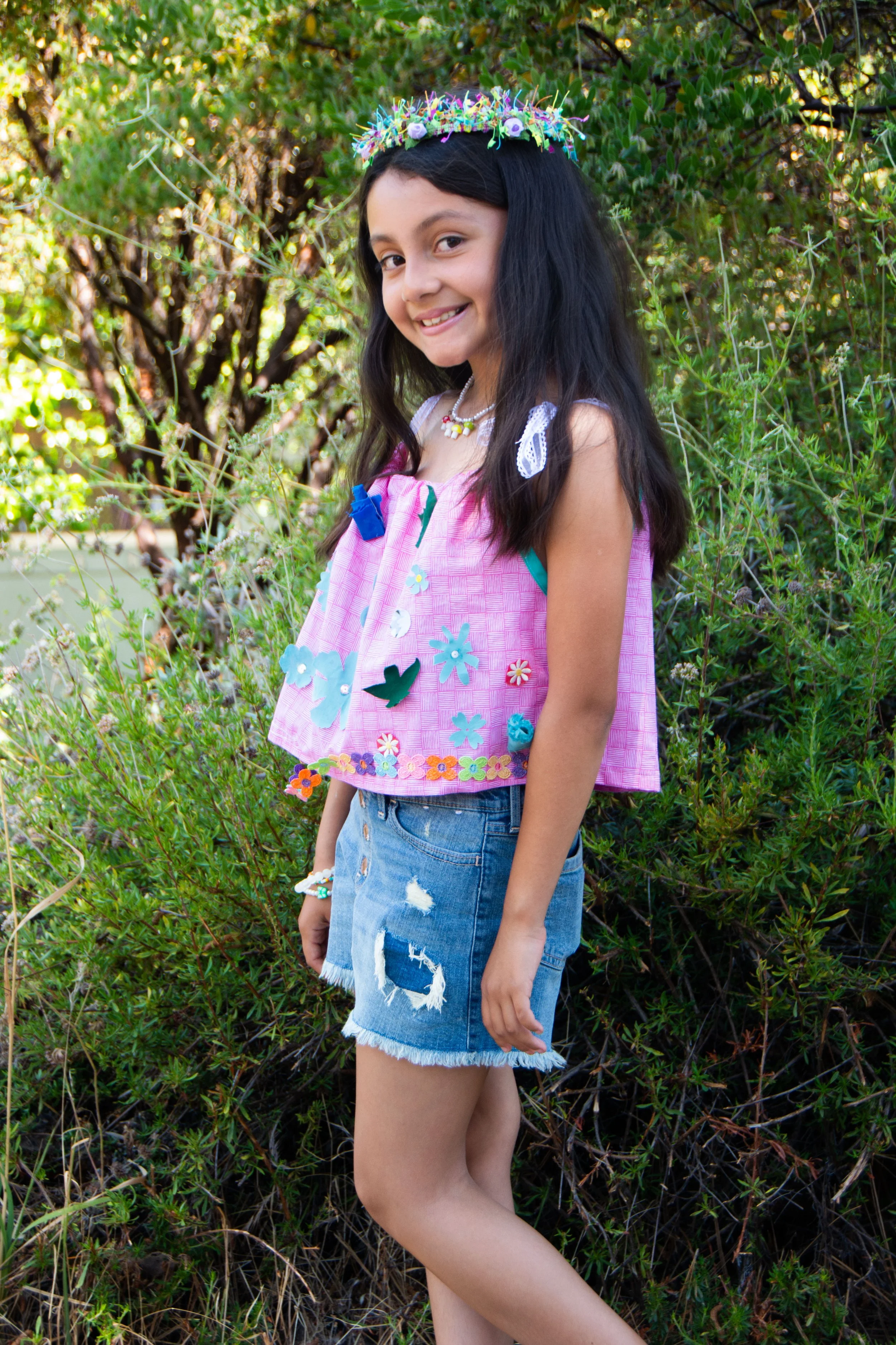 Smiling girl with long dark hair wearing a pink top with colorful butterflies and flowers, denim shorts, and a floral headband, standing outdoors among greenery.