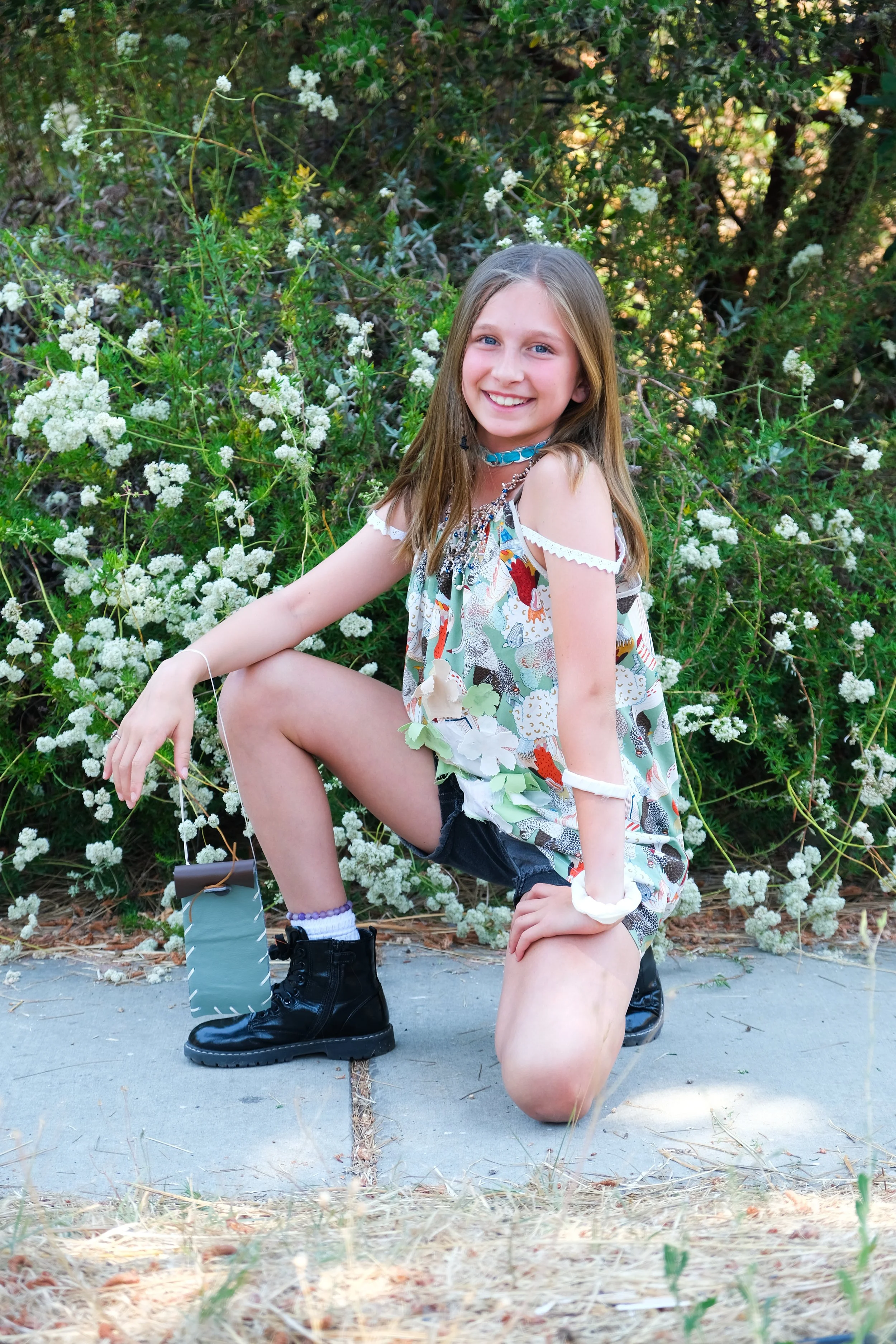 A young girl with long blonde hair and a colorful patterned top, wearing black shorts, black boots, a blue choker, and a white bracelet, is kneeling on one knee on a concrete sidewalk with greenery and white flowers in the background, smiling at the 