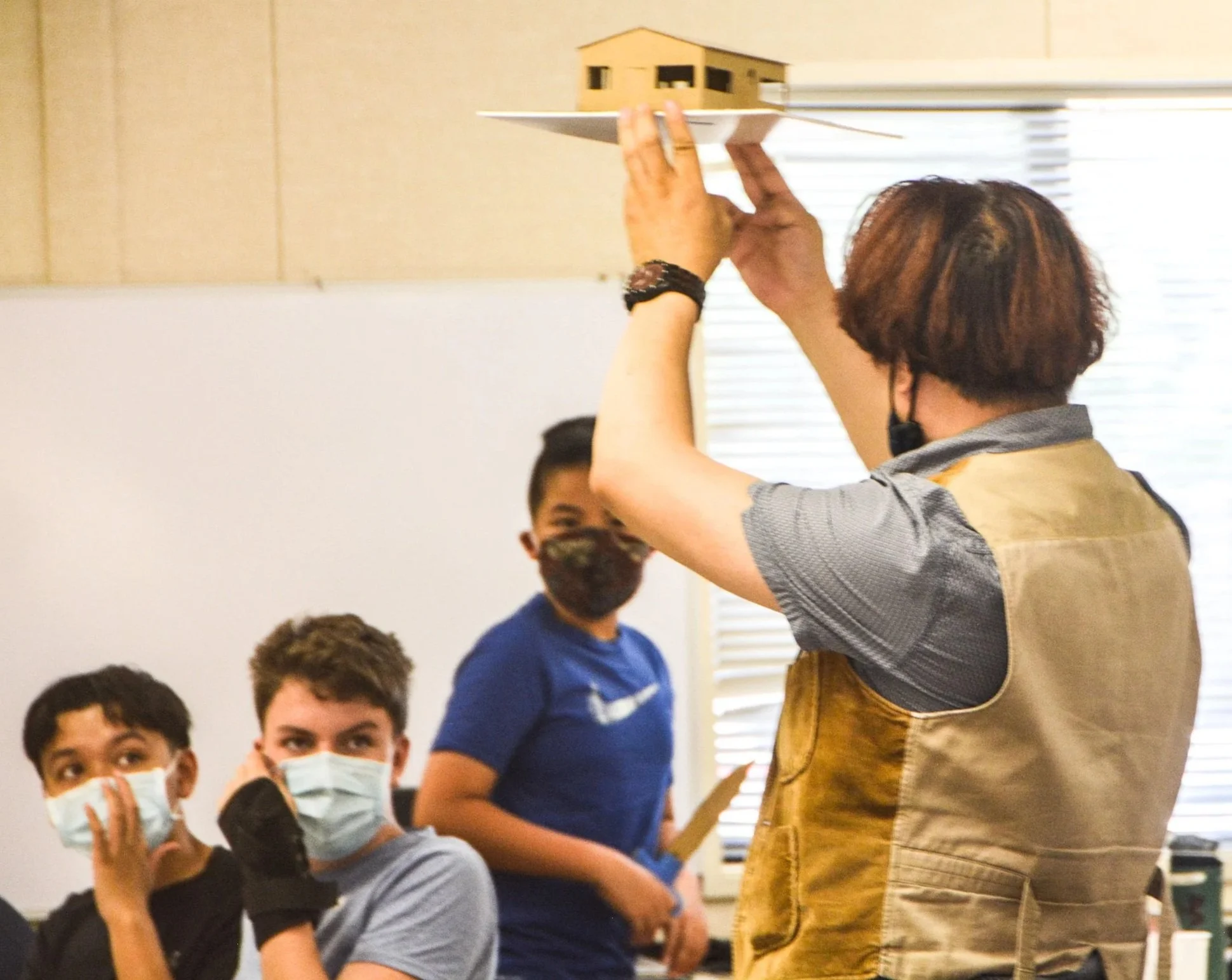 Woman holding a scale model of a house in a classroom with children wearing masks.