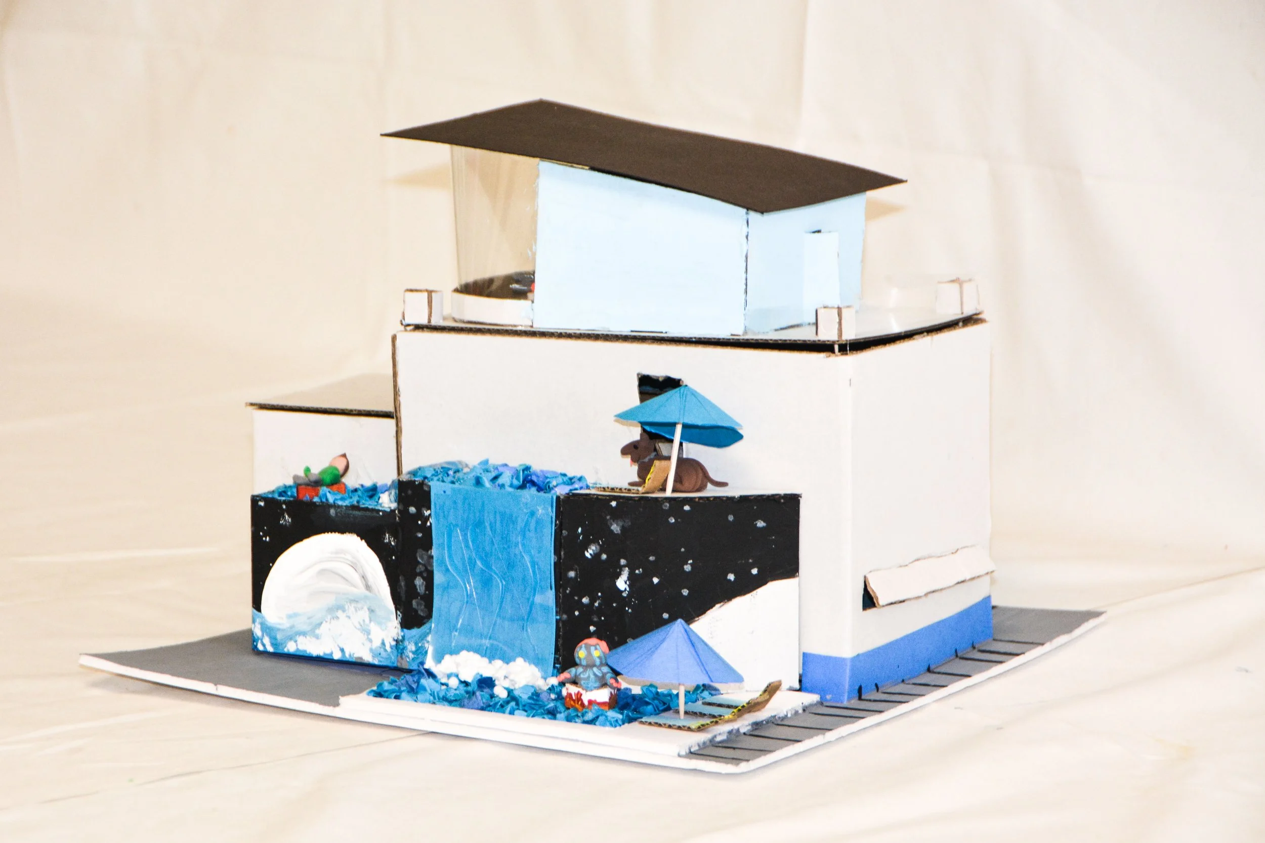 A handmade model of a modern, multi-level house with beach and ocean themed decorations, including umbrellas, a waterfall, and small beach scene elements.